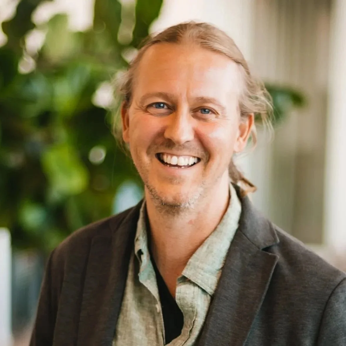 En man i en kavaj håller i ett par glasögon och ler mot kameran. I bakgrunden syns en grön växt och en soffa.