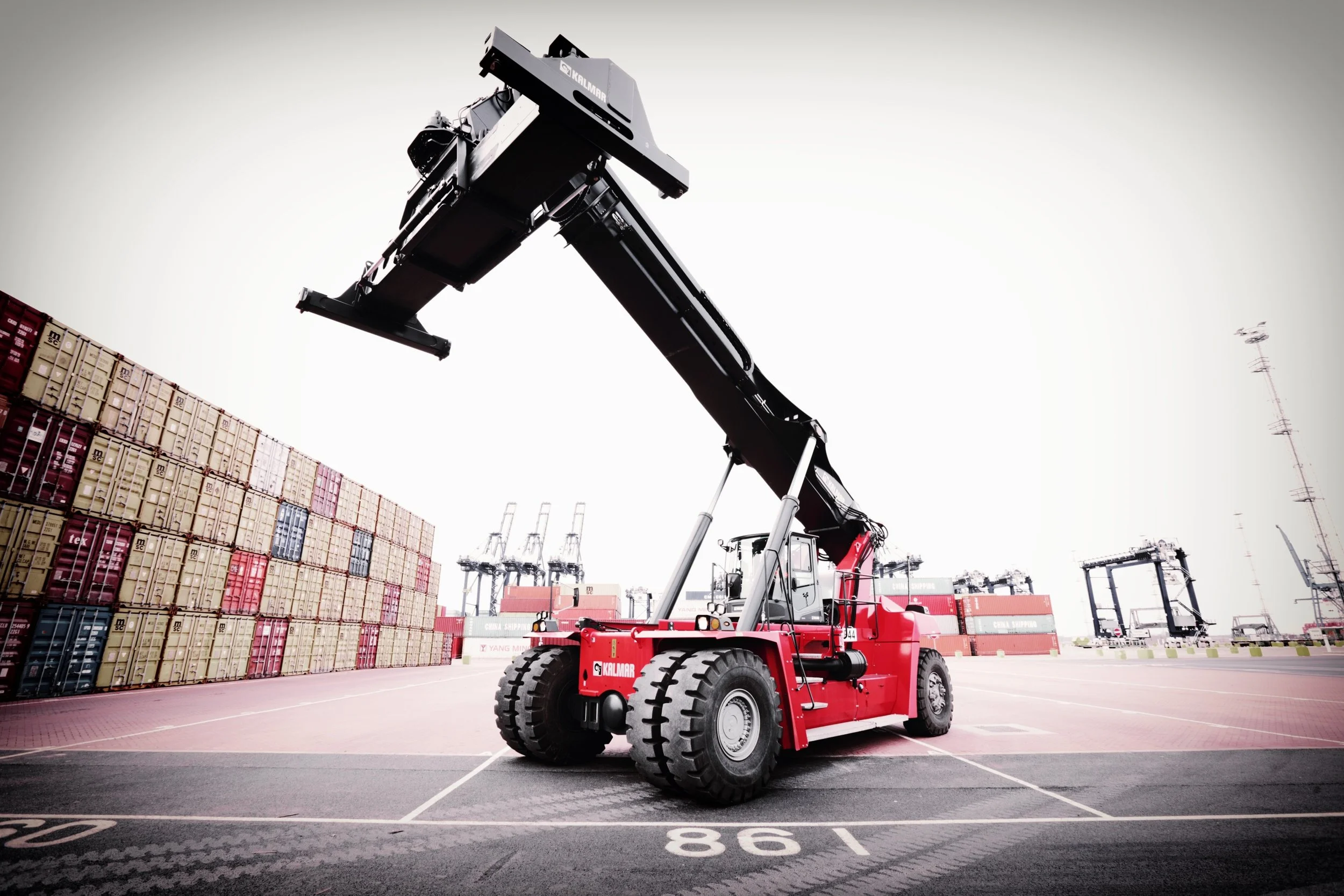 Reachstacker i hamnen som hanterar fraktcontainrar, med staplar av containrar i bakgrunden och kranar i fjärran.