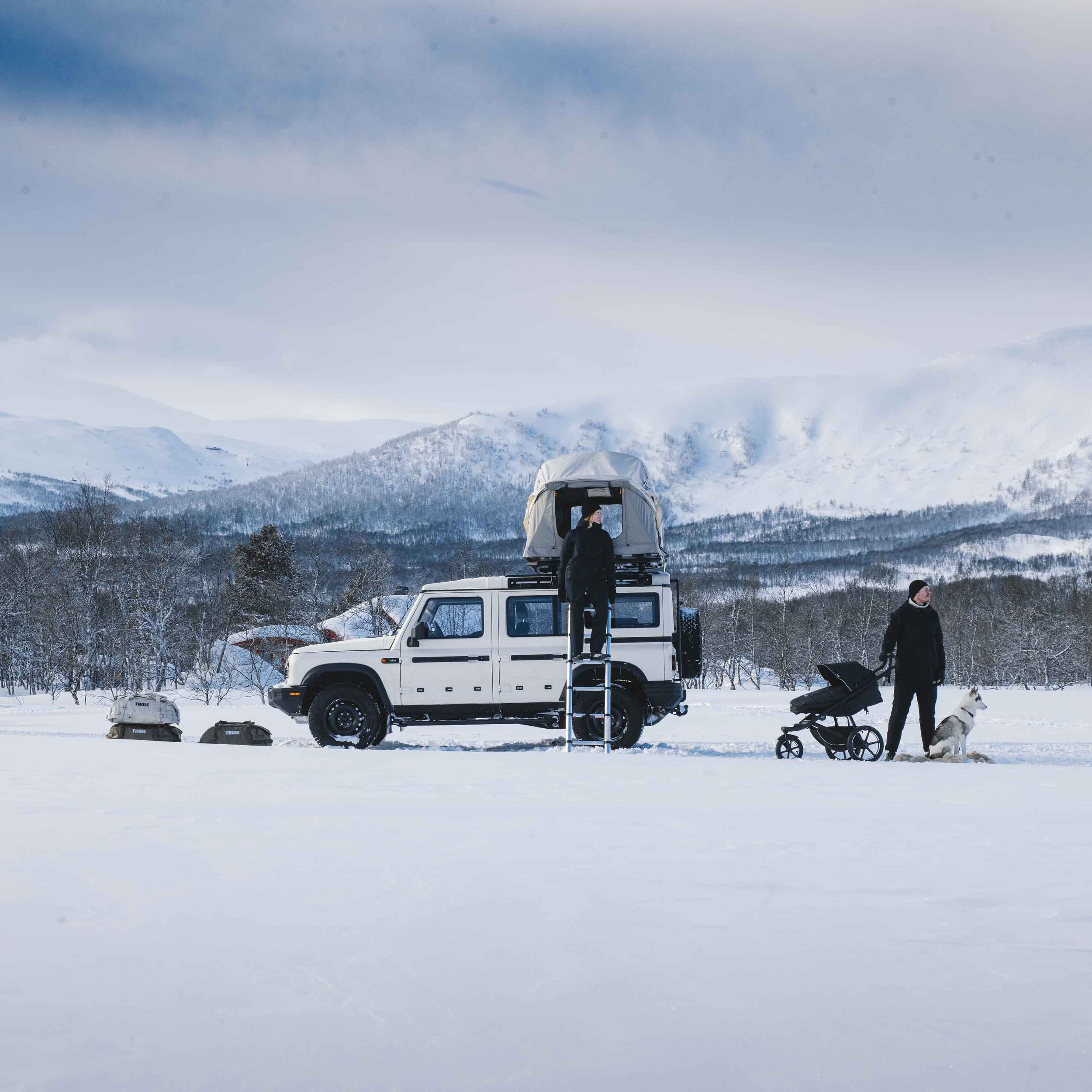 Vit terrängbil utrustad med taktält och stege från Thule i ett snöigt bergslandskap. En kvinna klättrar upp på stegen. Till höger står en man med en barnvagn och en hund. Bilden illustrerar Avalons kompetens inom innovativ produktutveckling.