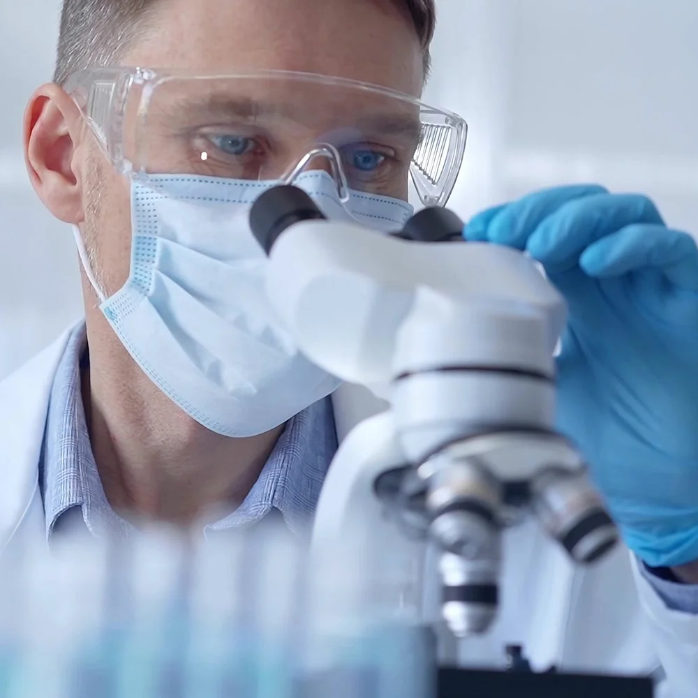 Forskare med skyddsglasögon och ansiktsmask använder mikroskop i laboratorium.
