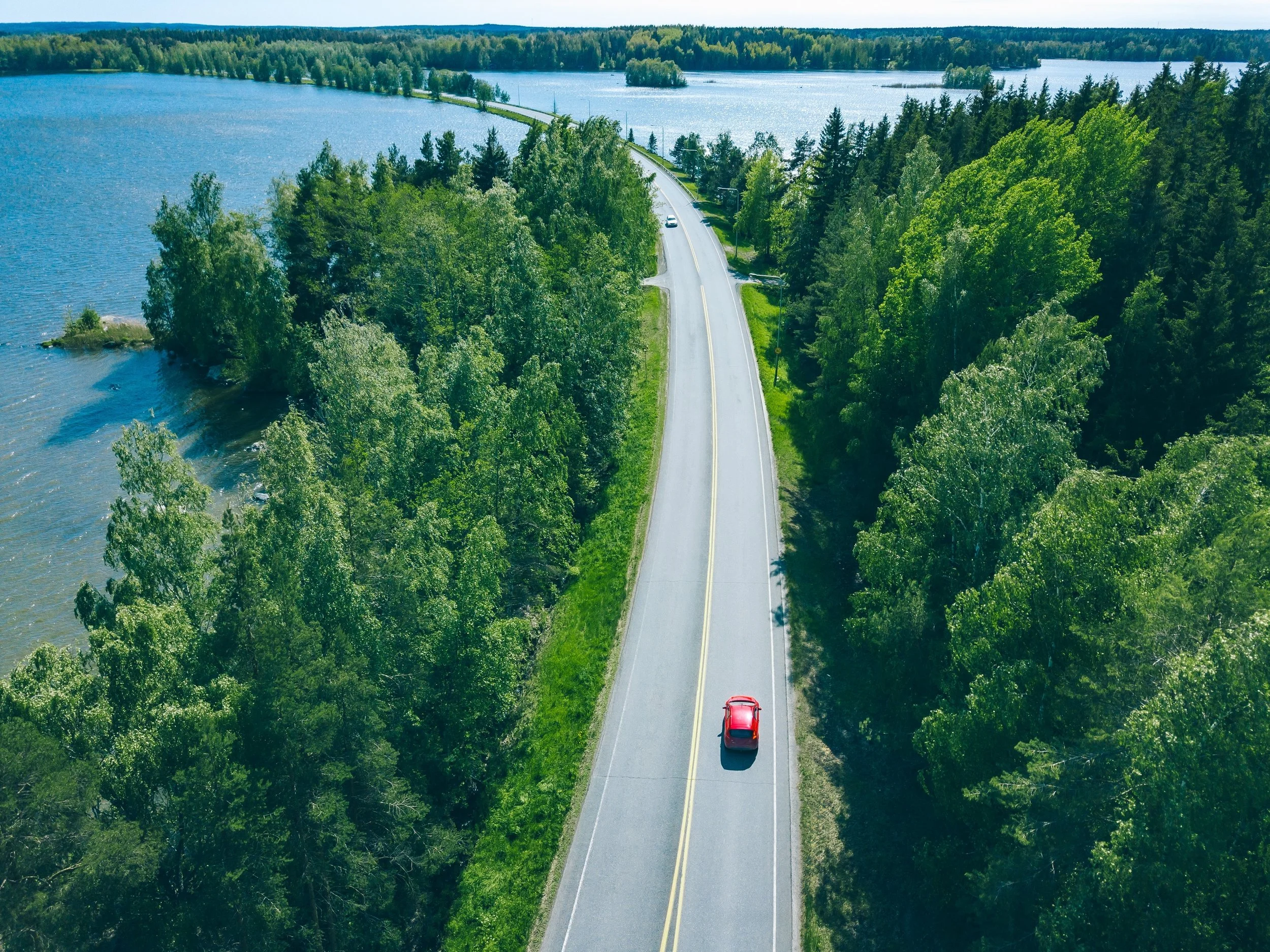 En röd bil kör på en vacker väg genom lövskog och vattendrag