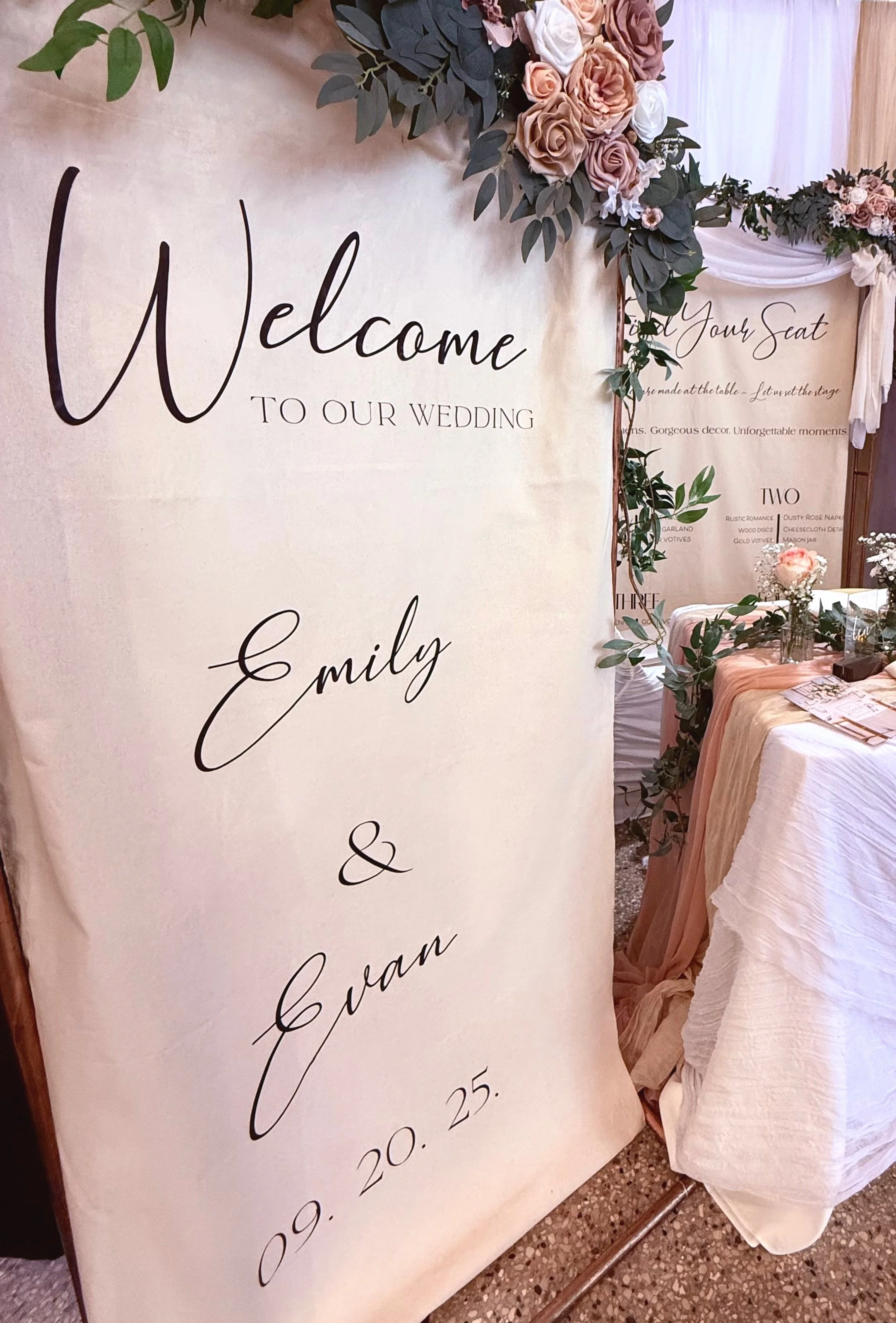 Wedding welcome sign with floral decor and table setting, featuring names "Emily & Evan" and date "09.20.25."
