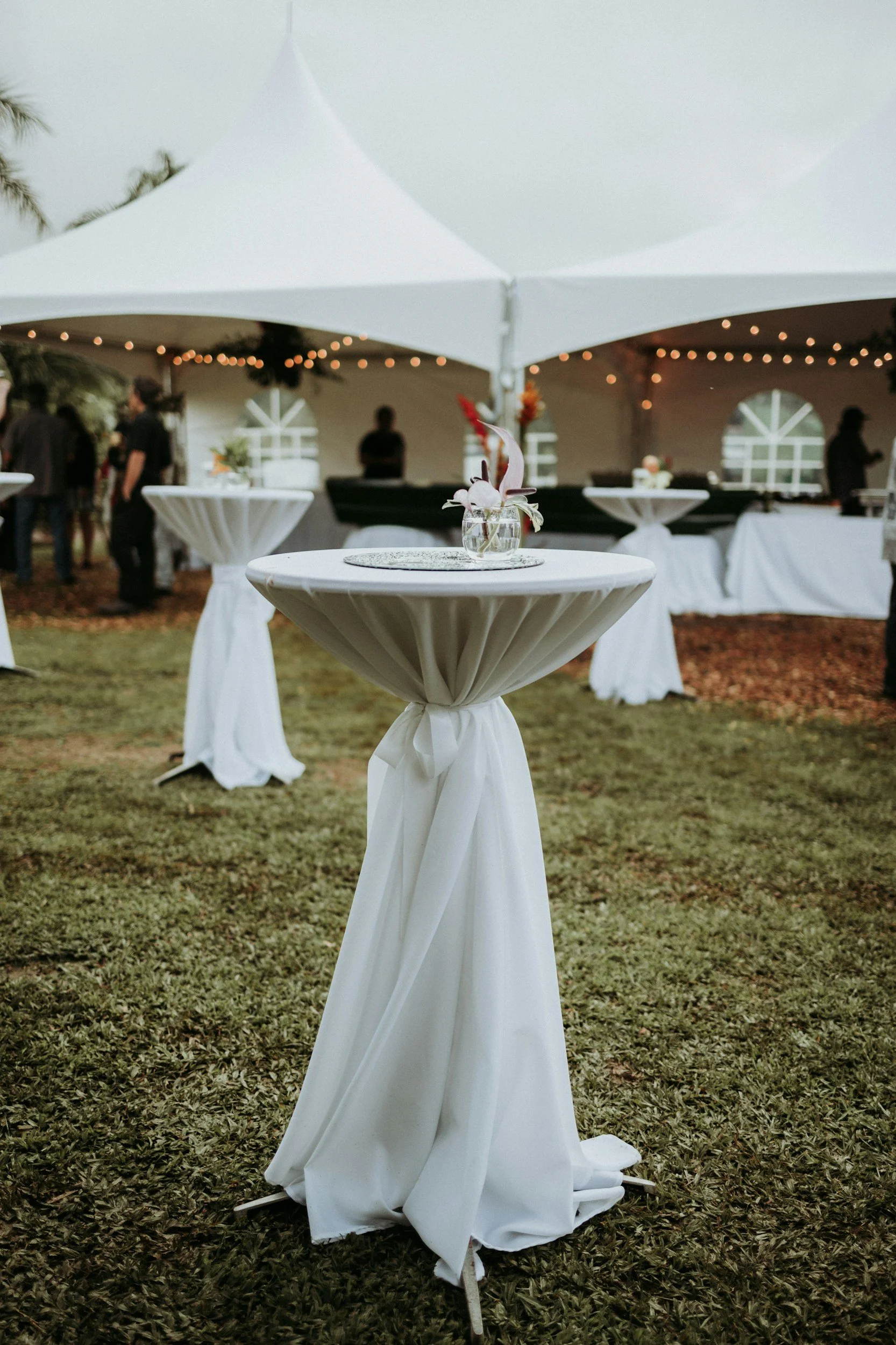 Outdoor event setup with white tables covered in cloth, string lights, and a white tent in the background.