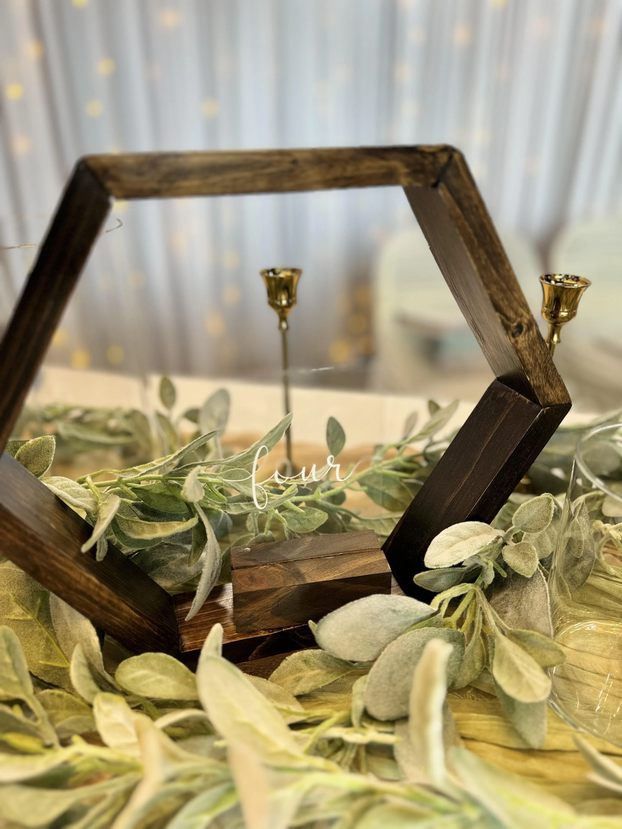 Wooden hexagon table centerpiece with glass inscribed "four," adorned with greenery and gold candle holders.