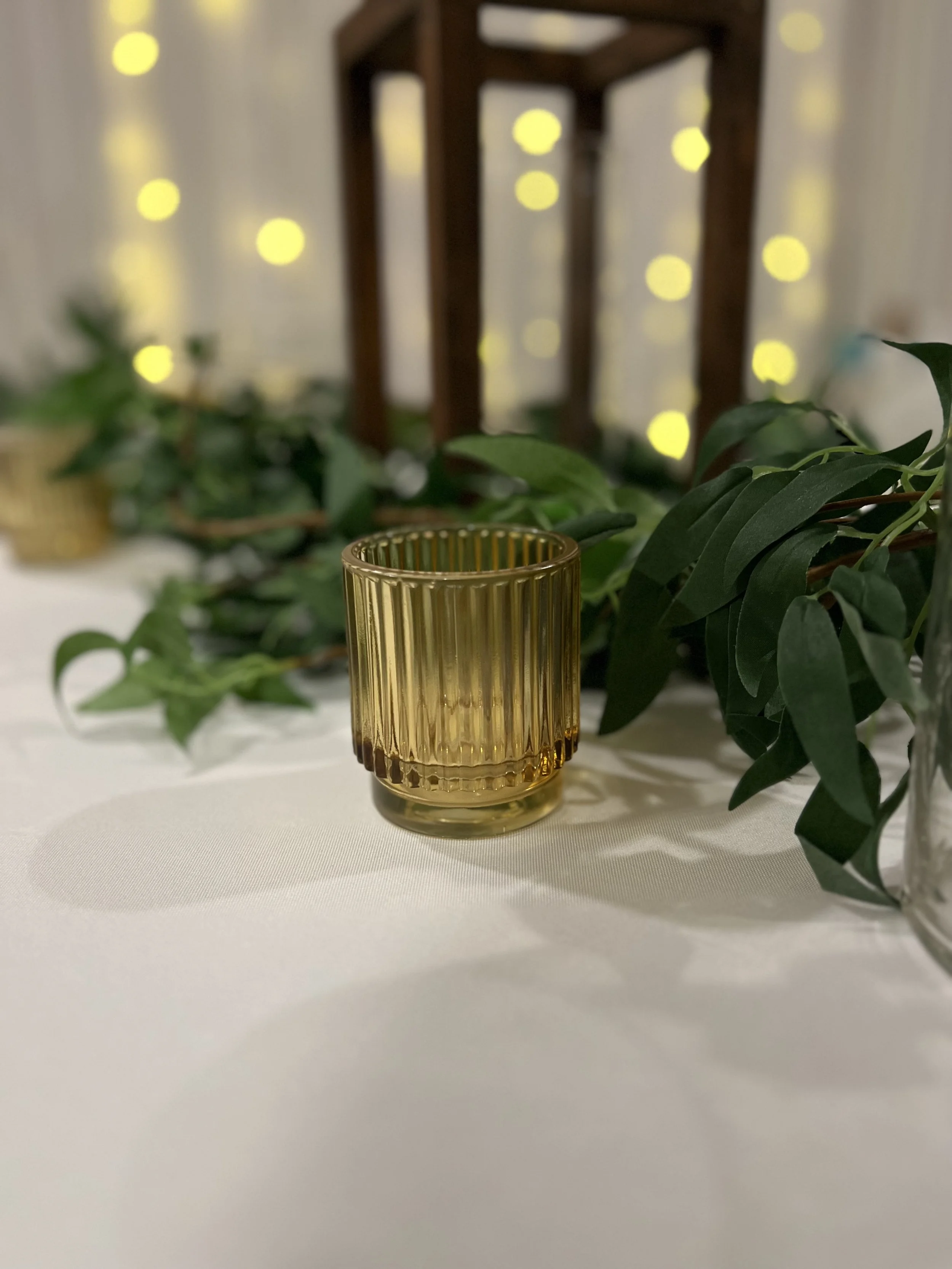 A ribbed amber glass candle holder placed on a table with greenery and a wooden lantern in the background, set against a backdrop of blurred, warm yellow lights.