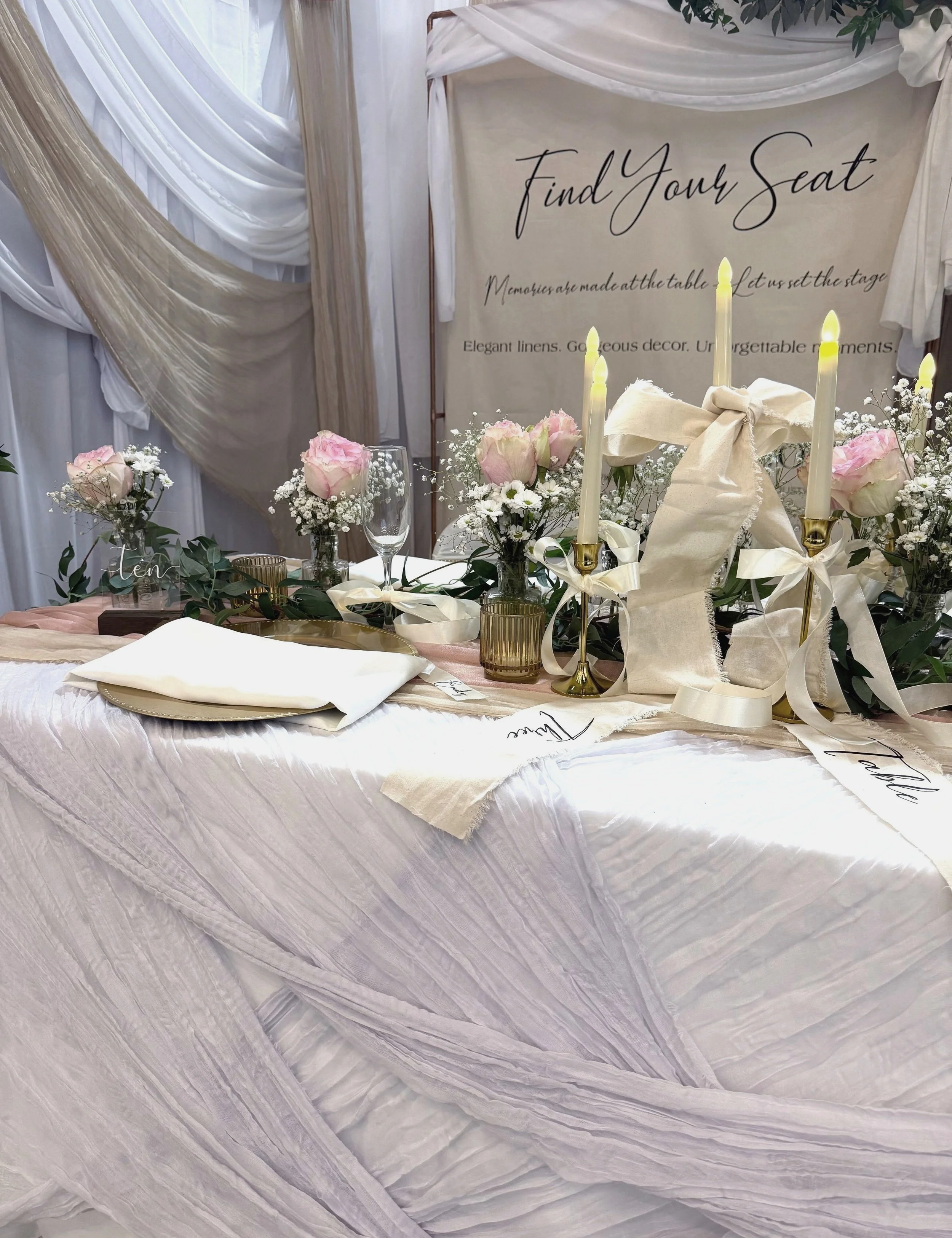Elegant wedding reception setup with draped fabric, pink roses, and candles. A sign saying "Find Your Seat" is in the background.