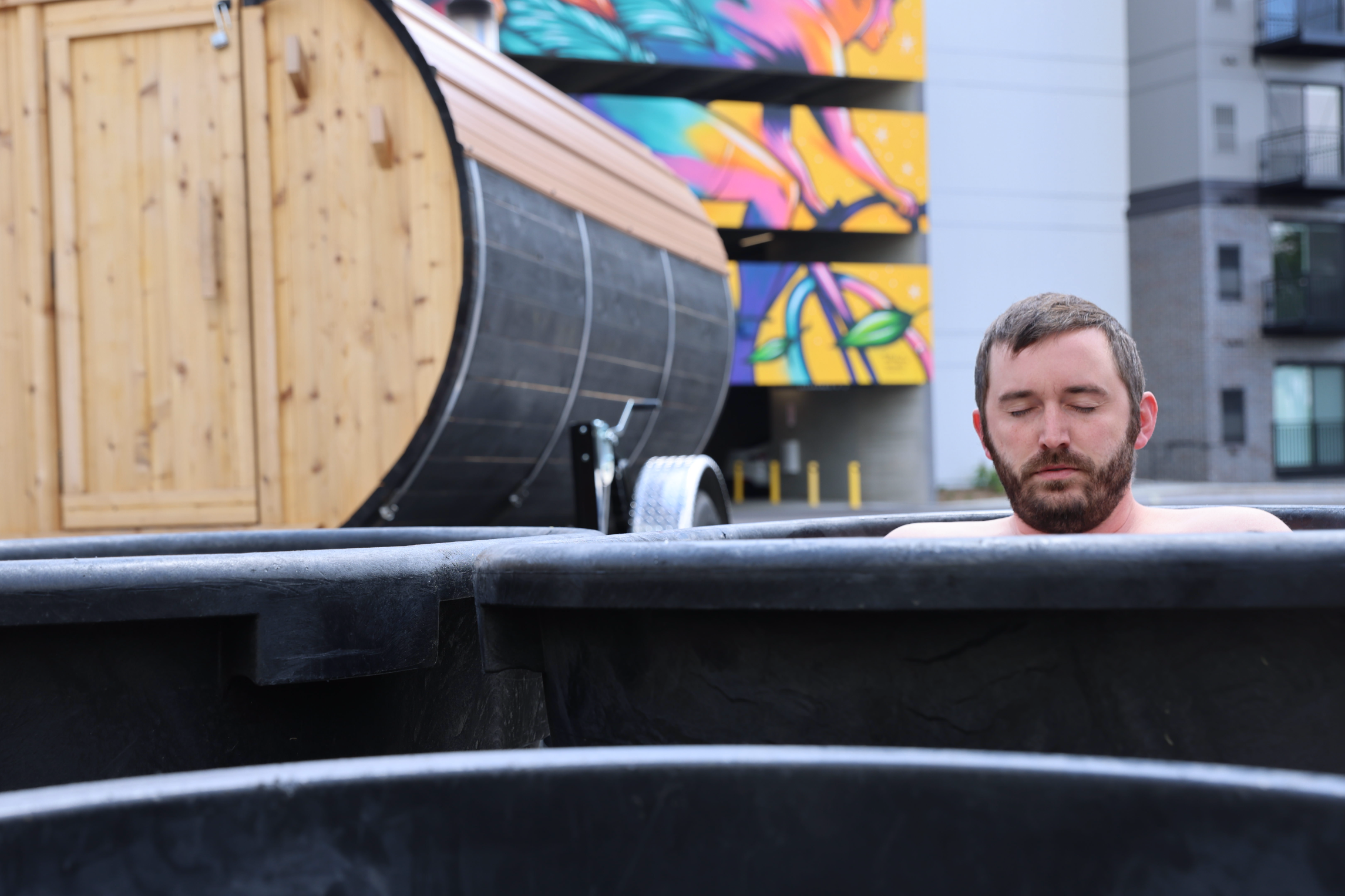 A man with a beard and closed eyes soaking in a black outdoor tub, with colorful graffiti and urban buildings in the background.