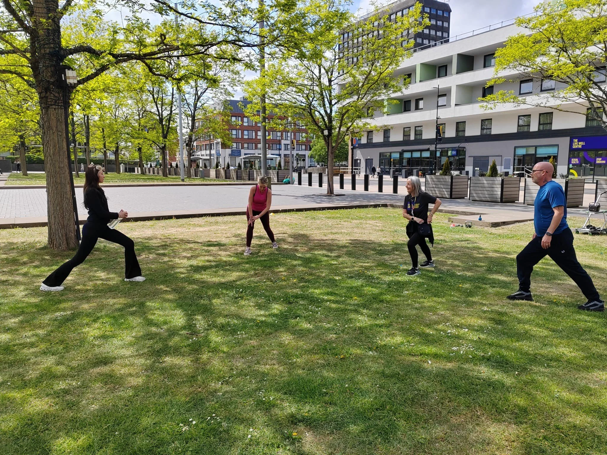 Four people practicing Tai Chi or a similar exercise on a grassy park area with trees and buildings in the background.