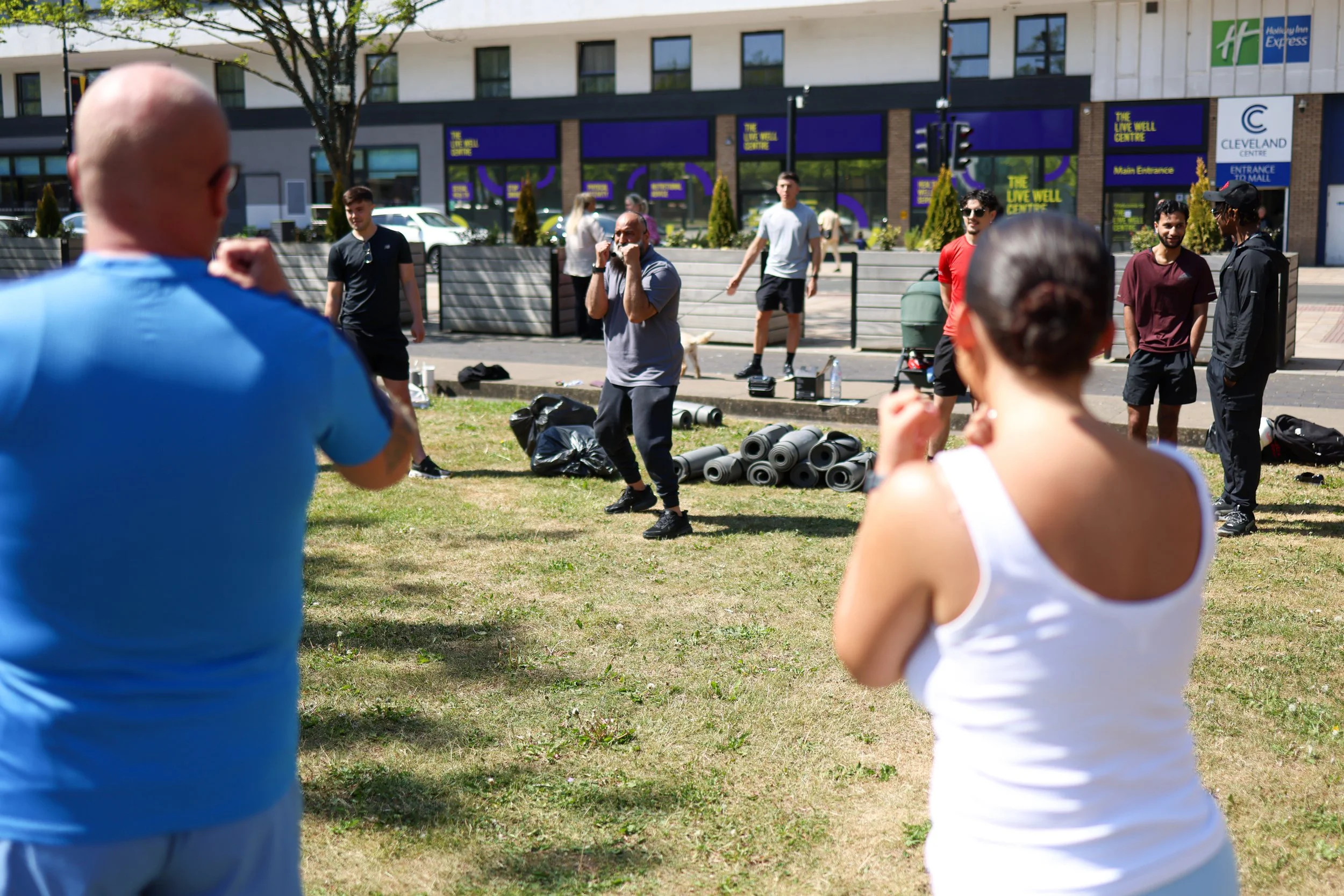 A group of people participating in an outdoor fitness class or workout session on a grassy area in a city, with some standing and others performing exercises, and fitness equipment such as yoga mats and water bottles visible on the ground.