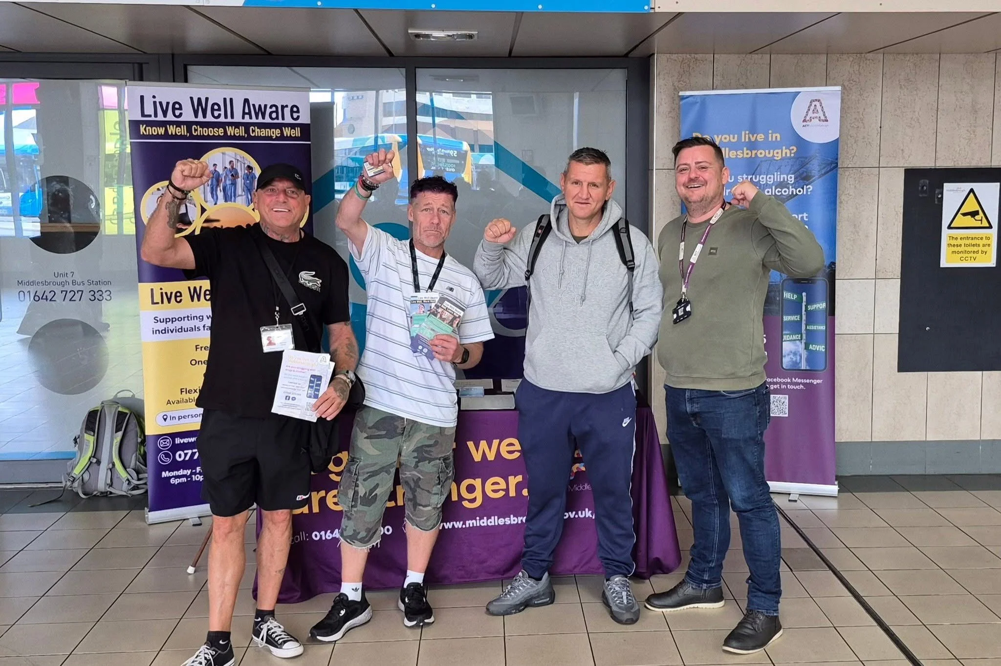 Four men standing inside a bus station, raising their fists. They are in front of a table with a purple tablecloth and banners promoting mental health awareness and support. Each person is wearing casual clothing and has a lanyard with an ID badge.