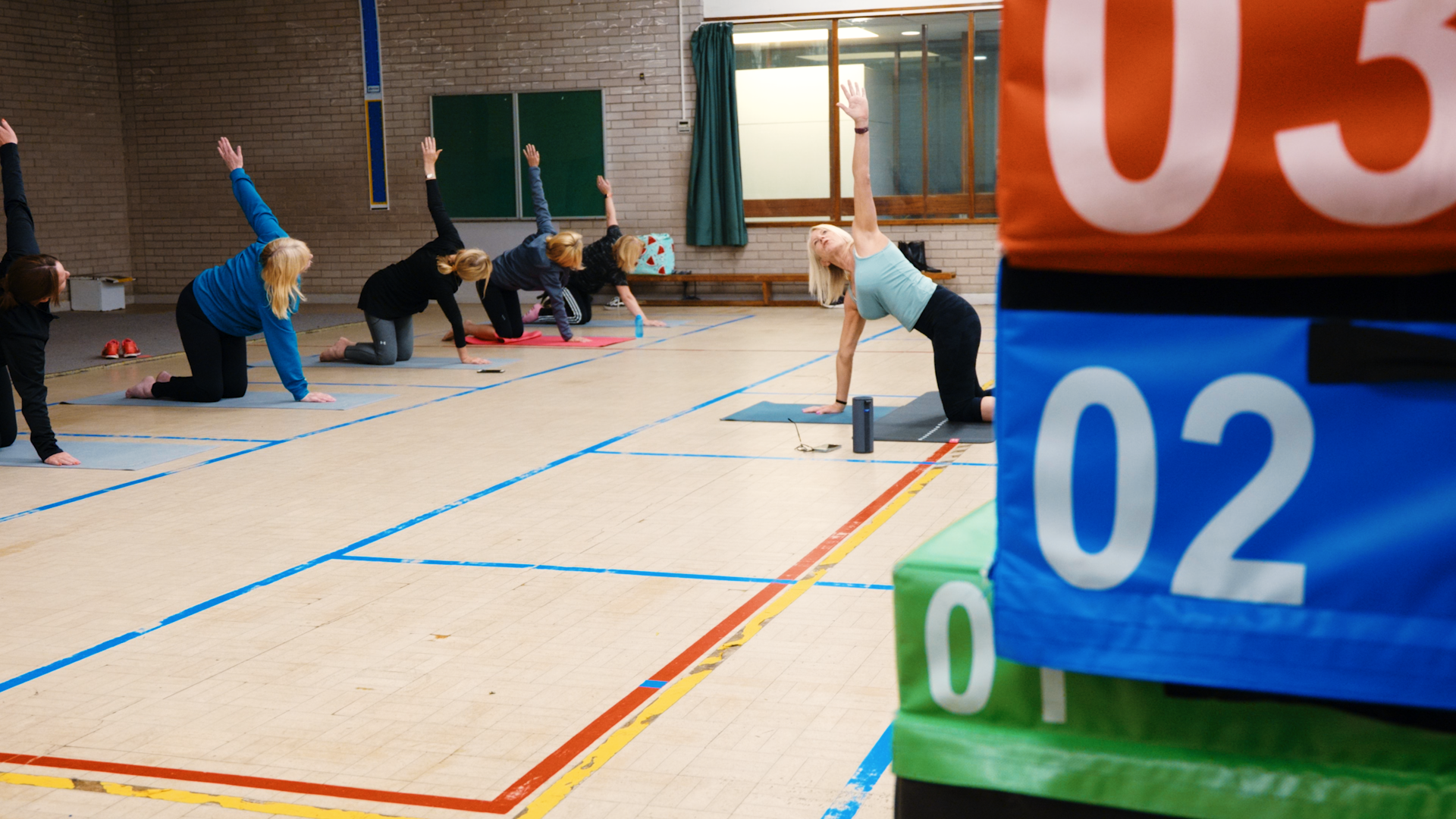 Group of women doing yoga on mats in a gymnasium, guided by an instructor.