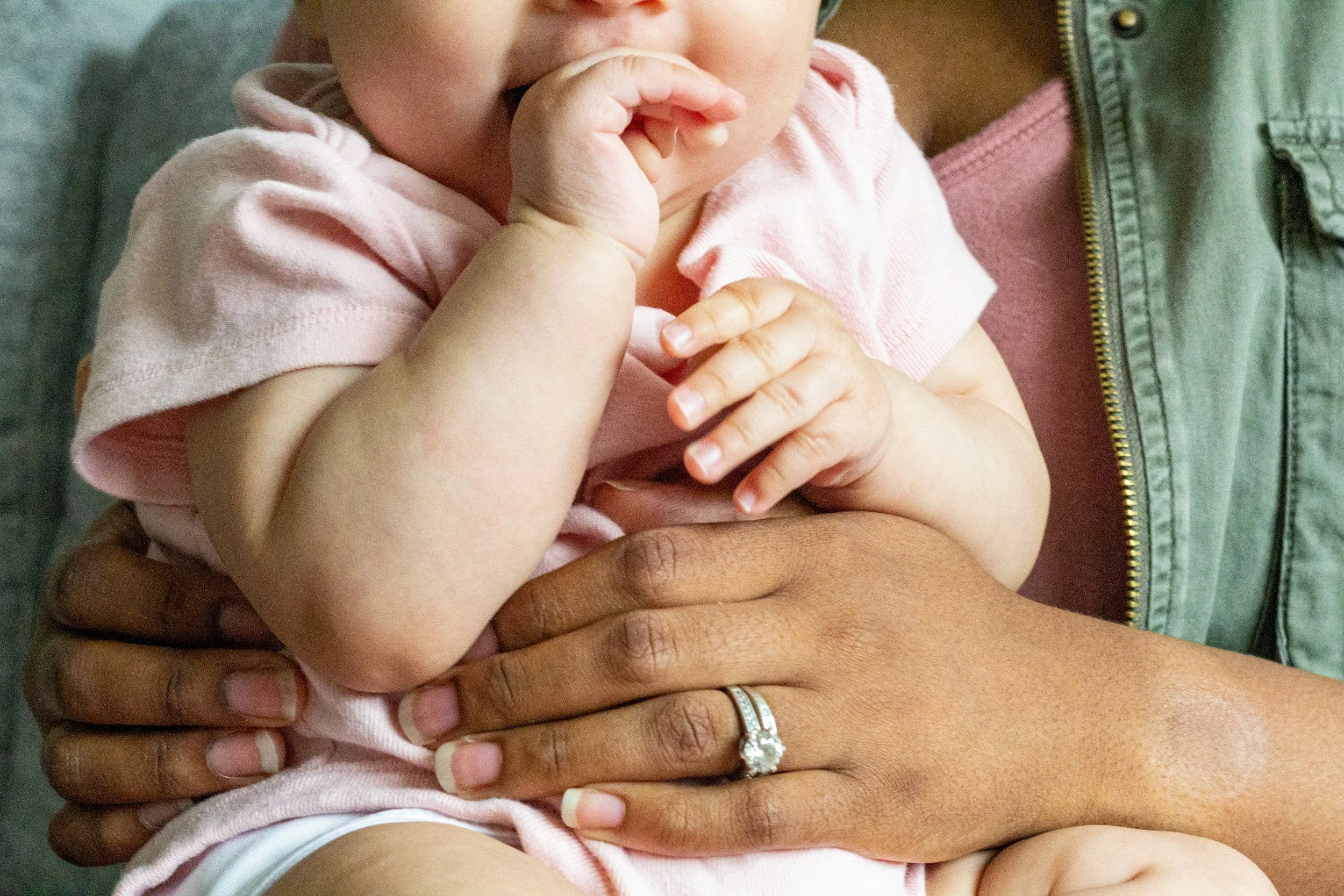 Close-up of a baby being held, with a focus on the baby's hand near their mouth, and an adult's hand with a wedding ring supporting them.