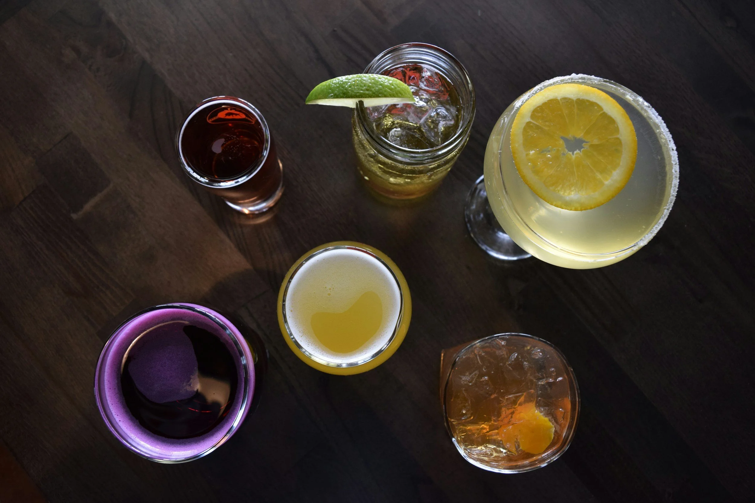 An overhead view of six different cocktails on a dark wooden table. The drinks include a dark red beverage, a lime and ice drink in a mason jar, a lemon slice in a glass of lemonade, a purple drink with ice, a cloudy yellow drink, and an amber-colored drink with a piece of citrus.