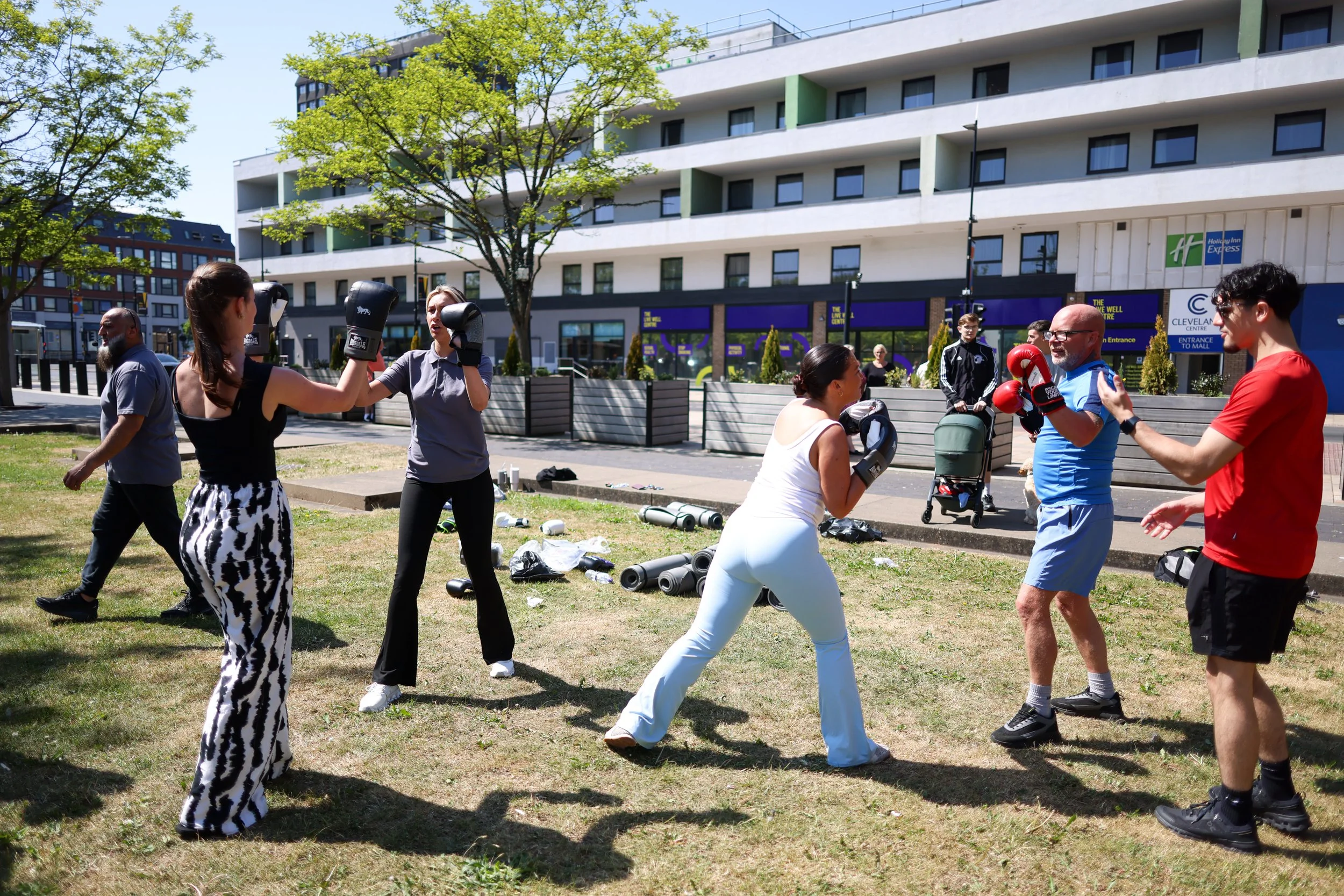 Group of people practicing boxing outdoors in a park, with some wearing gloves and focusing on each other, while others observe or prepare to spar, in front of an apartment building.