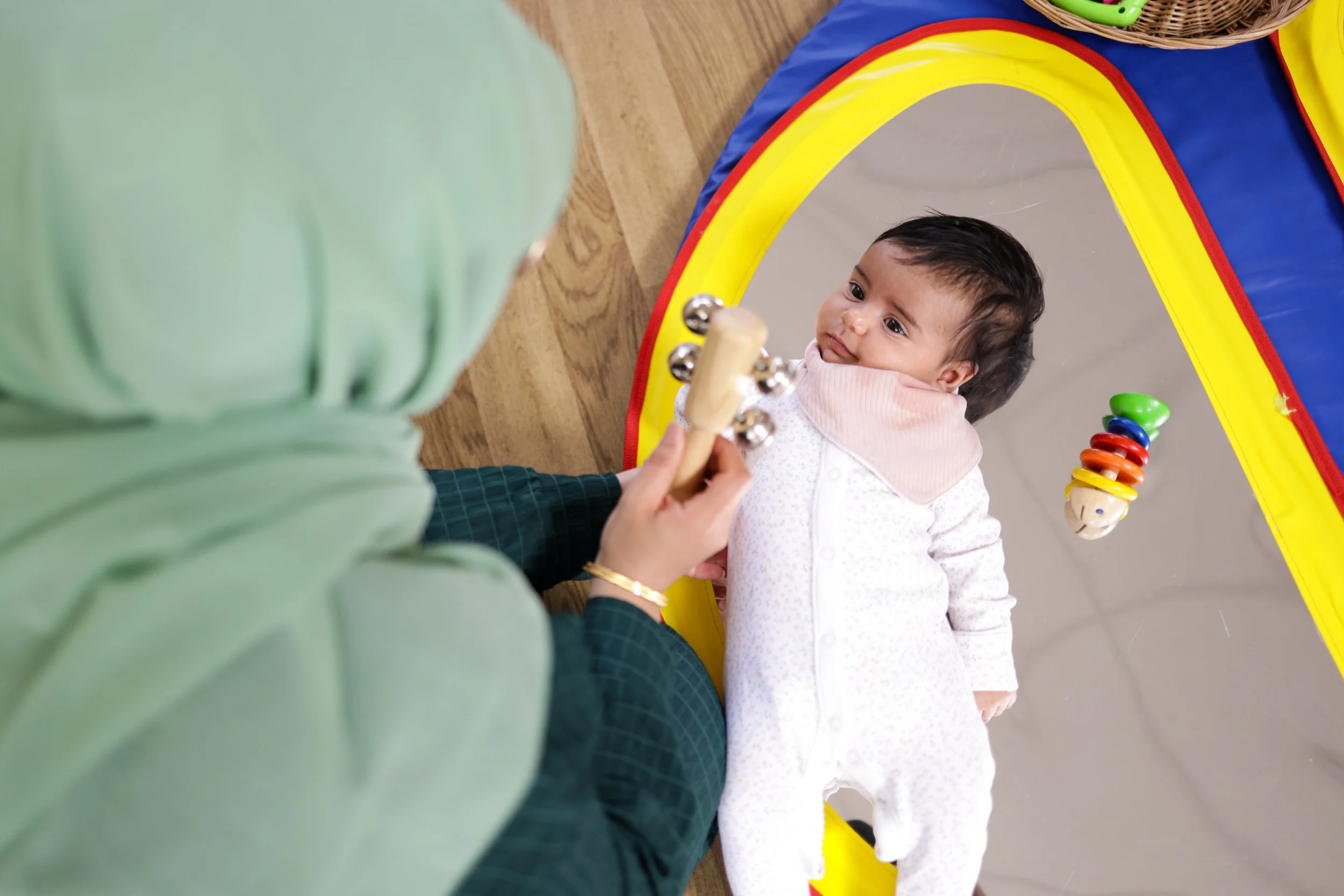 A woman in a green jacket playing with a baby lying on a colorful play mat.