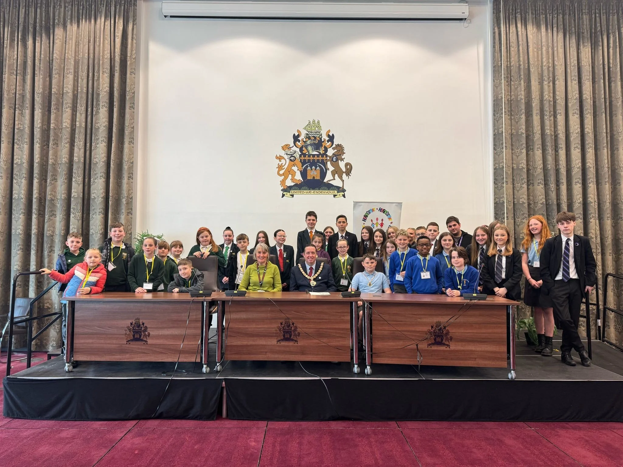 Group of children and adults posing for a photo in a conference room, with a large crest on the wall behind them and a banner reading "Happiness is Here".