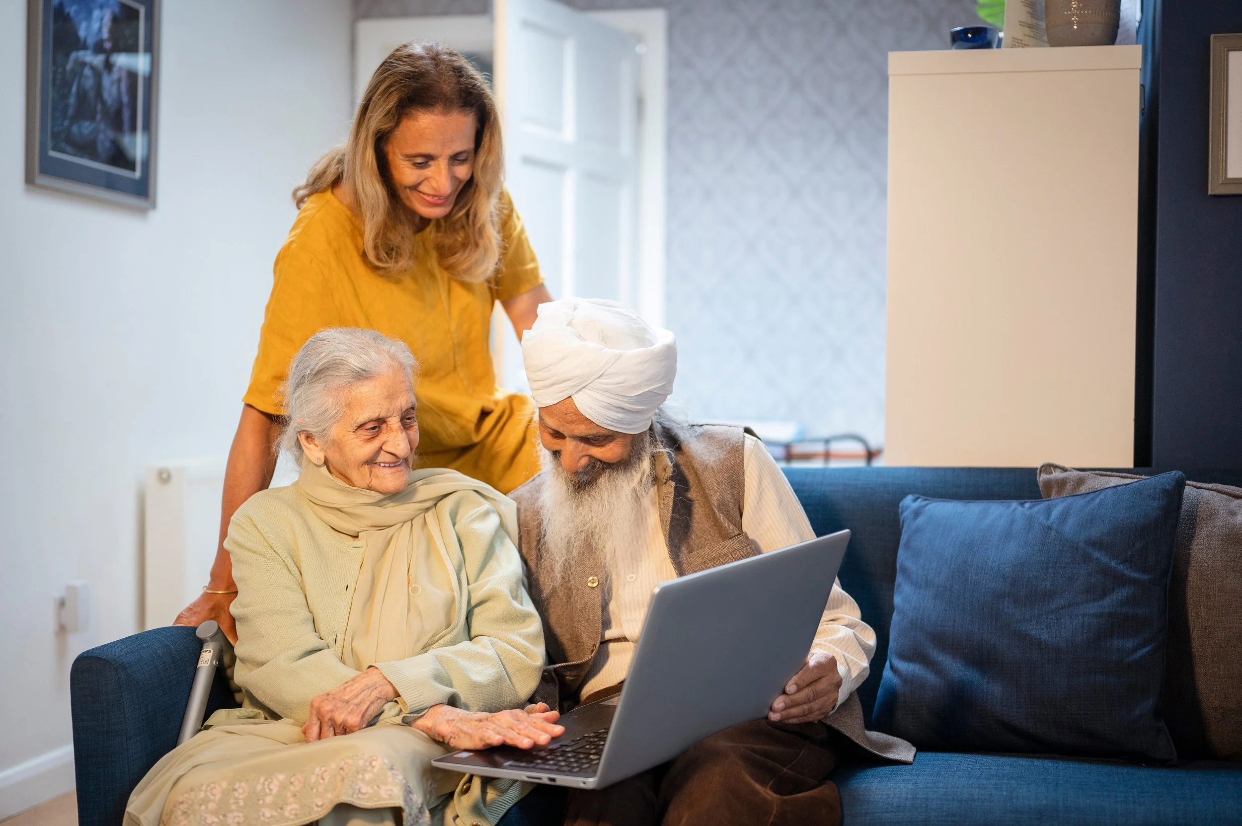 A group of four people, including an elderly woman with a cane, a man wearing a turban, a woman in a yellow top, and another woman, gathered around a laptop on a couch in a living room, looking at the screen and smiling.