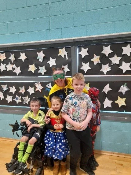 Group of children and an adult dressed in superhero costumes, standing in front of a classroom decorated with star-shaped cutouts on a blackboard.