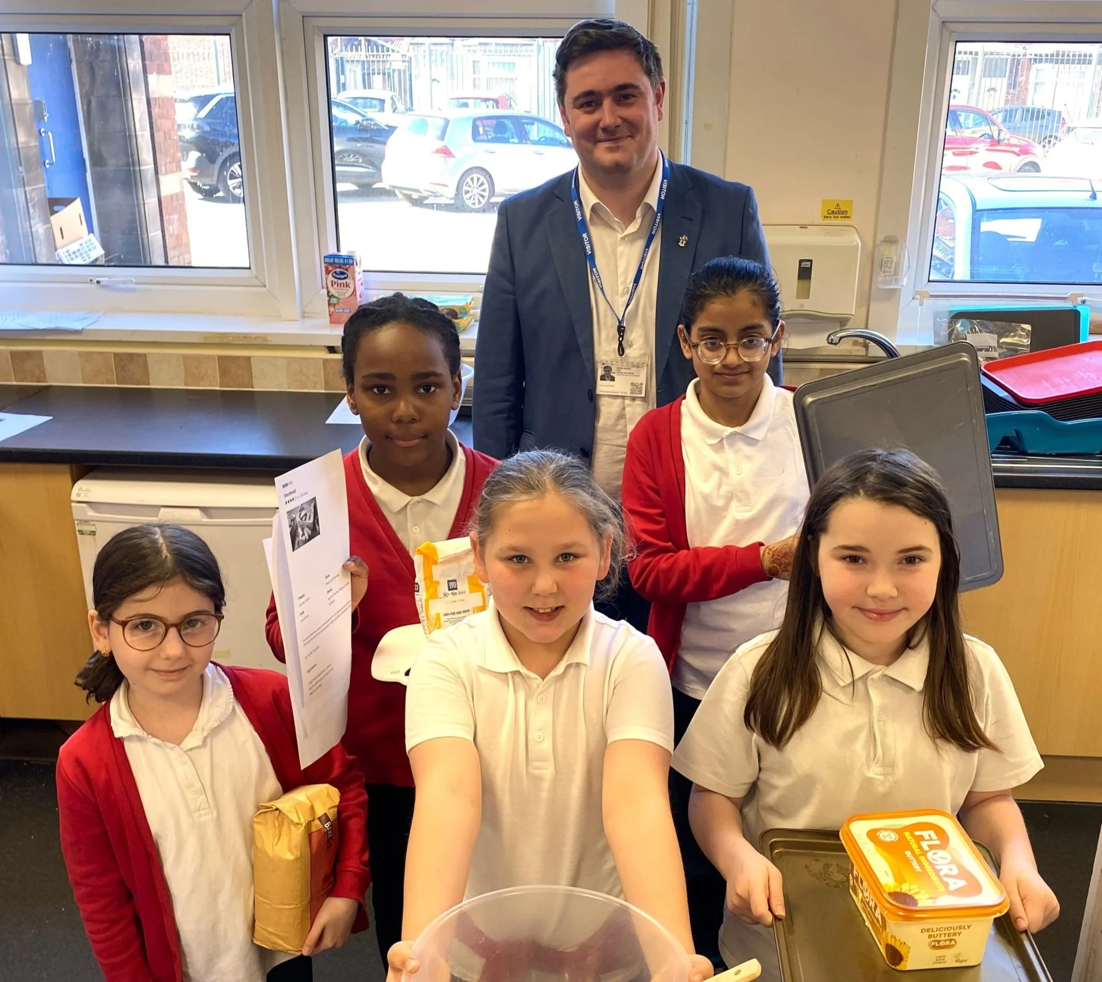 Group of five school children with an adult teacher in a classroom, some holding papers and food, posing for a group photo.