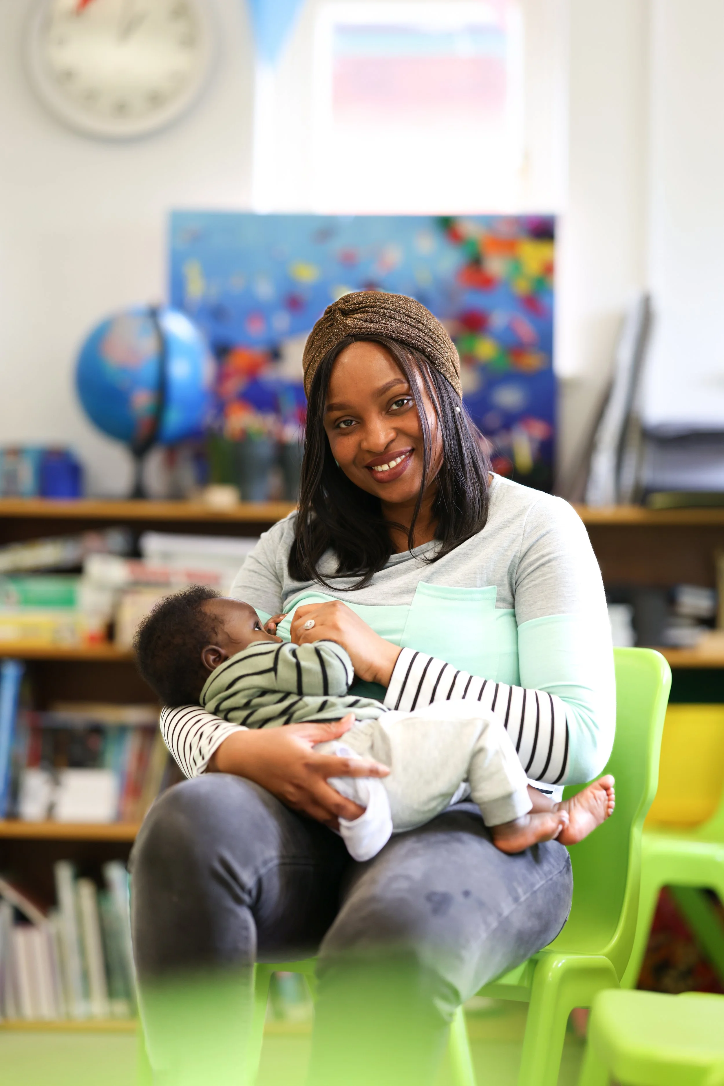 A woman breastfeeding a young child in a classroom or library setting, with bookshelves and colorful educational materials in the background.