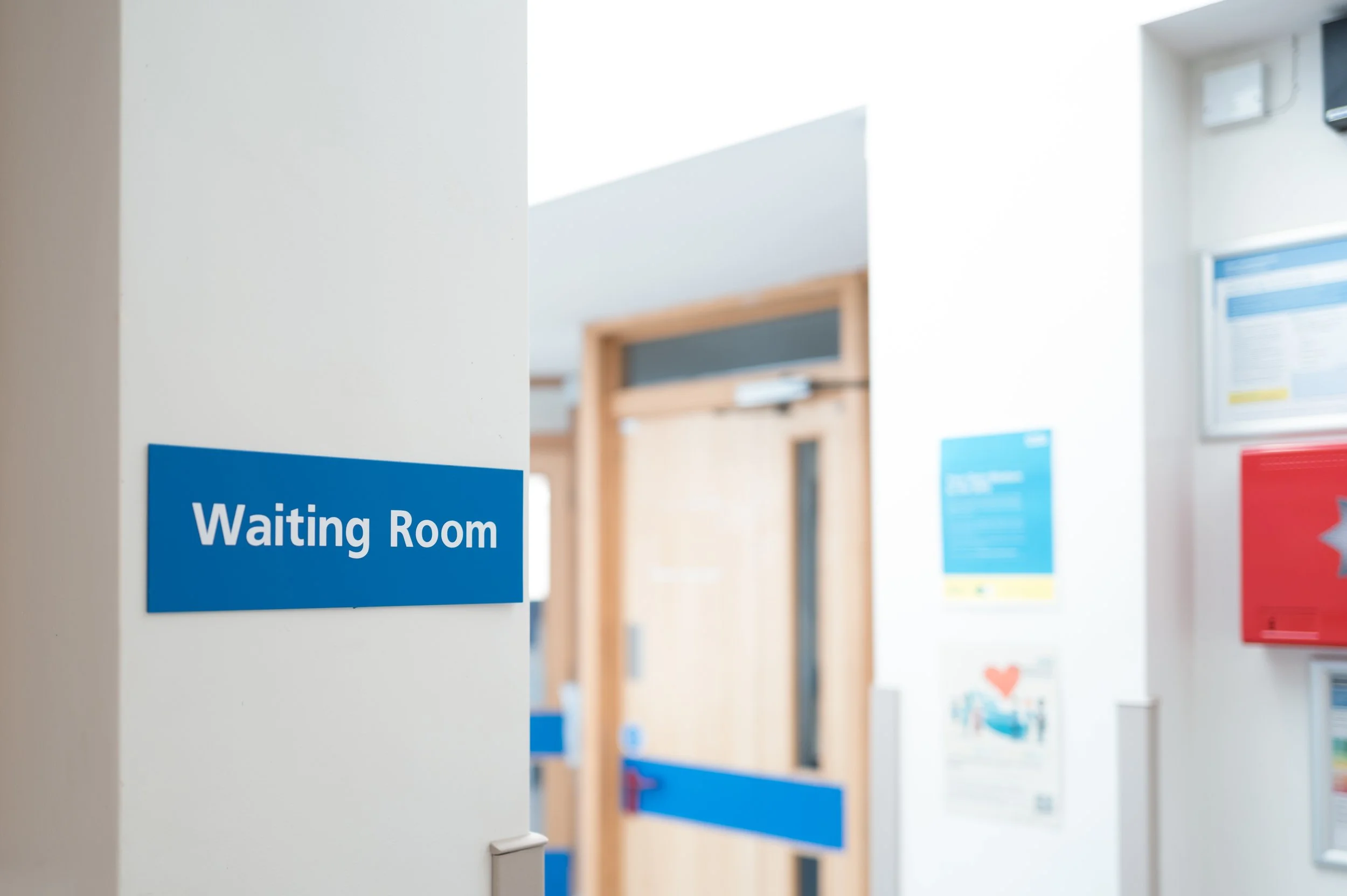 Indoor view of a hallway with a blue sign that says 'Waiting Room' on a white wall, with a wooden door in the background, and various informational posters on the walls.