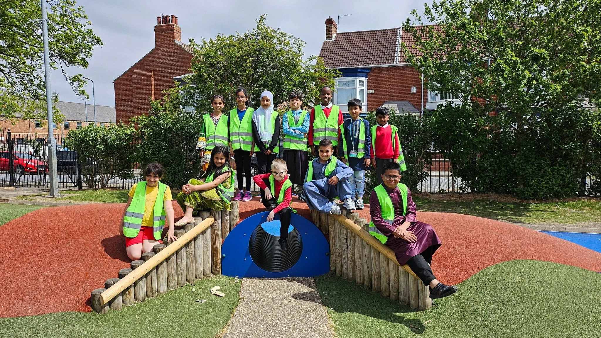 Group of children playing on a playground structure with a sloped wooden bridge and tunnel, surrounded by trees and houses, wearing bright safety vests.