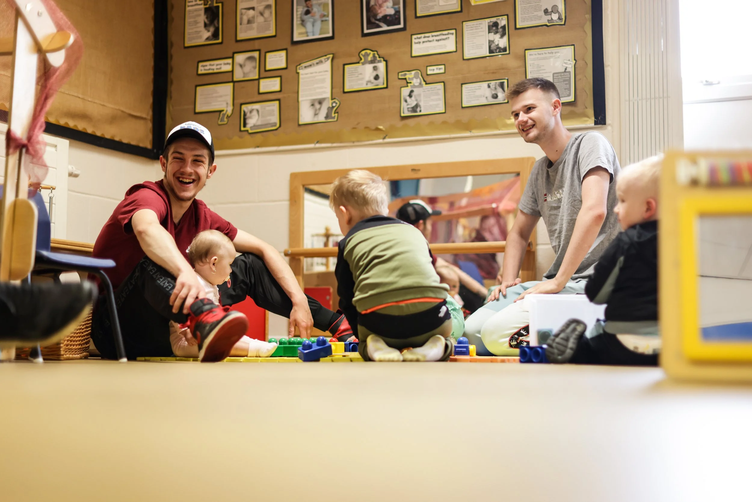 Two men and three young children sitting and playing with colorful toy blocks on the floor in a brightly lit room, likely a classroom or daycare, with a bulletin board on the wall behind them.