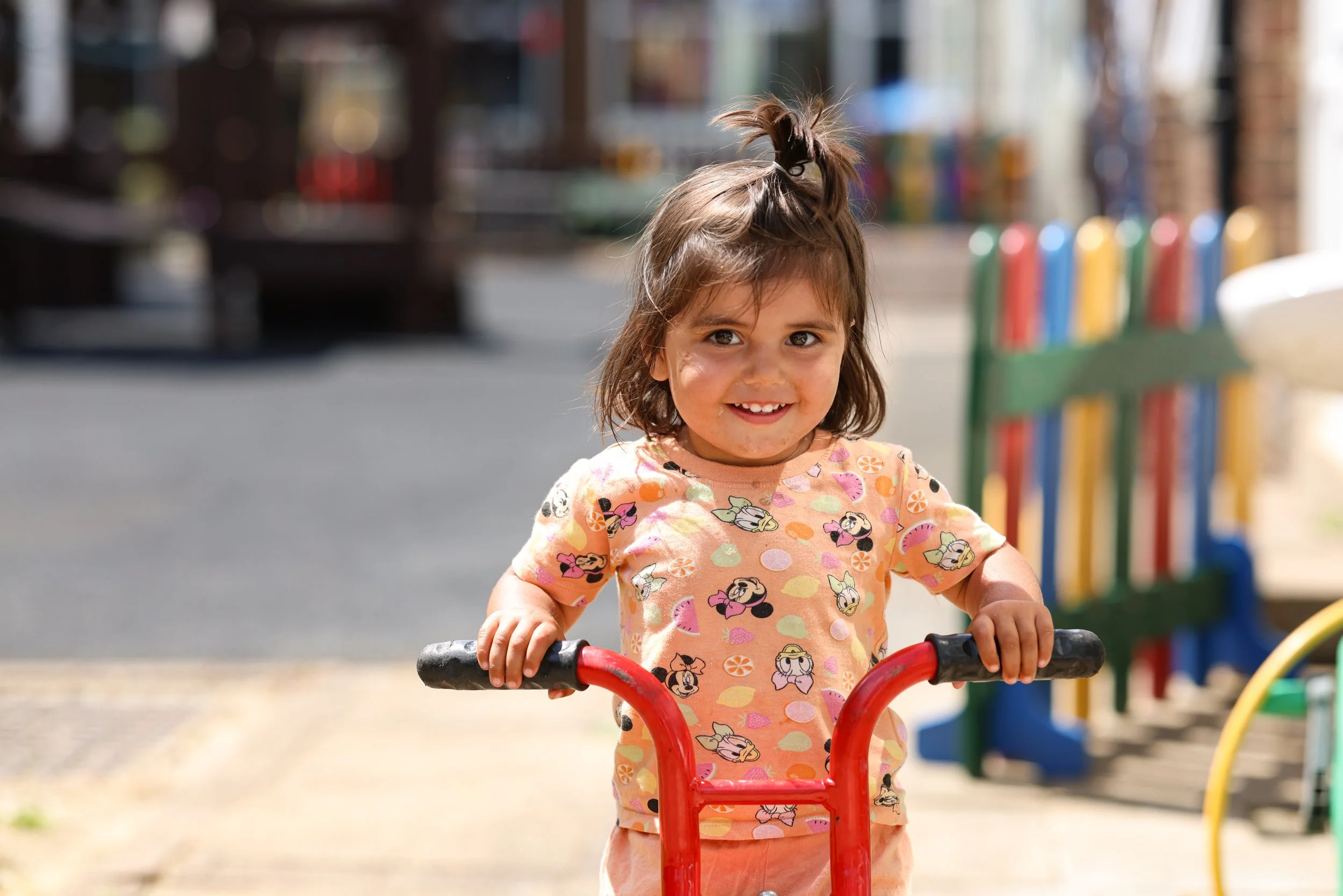 A young girl smiling and holding onto a small red scooter, standing outdoors in a park or playground area on a sunny day.