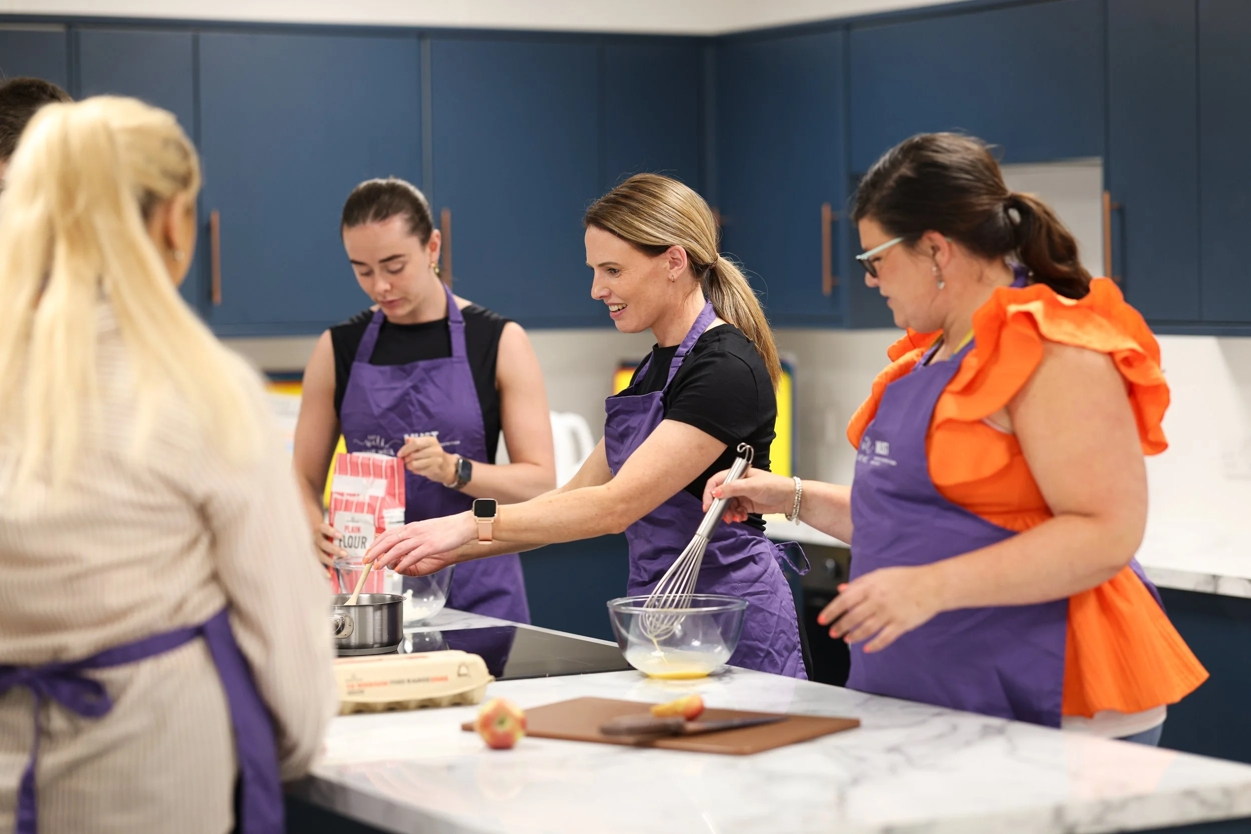 Four women wearing purple aprons are cooking together in a modern kitchen. One woman is pouring ingredients into a bowl, another is whisking in a bowl, and the others are assisting or observing. The kitchen has blue cabinets and a white countertop.