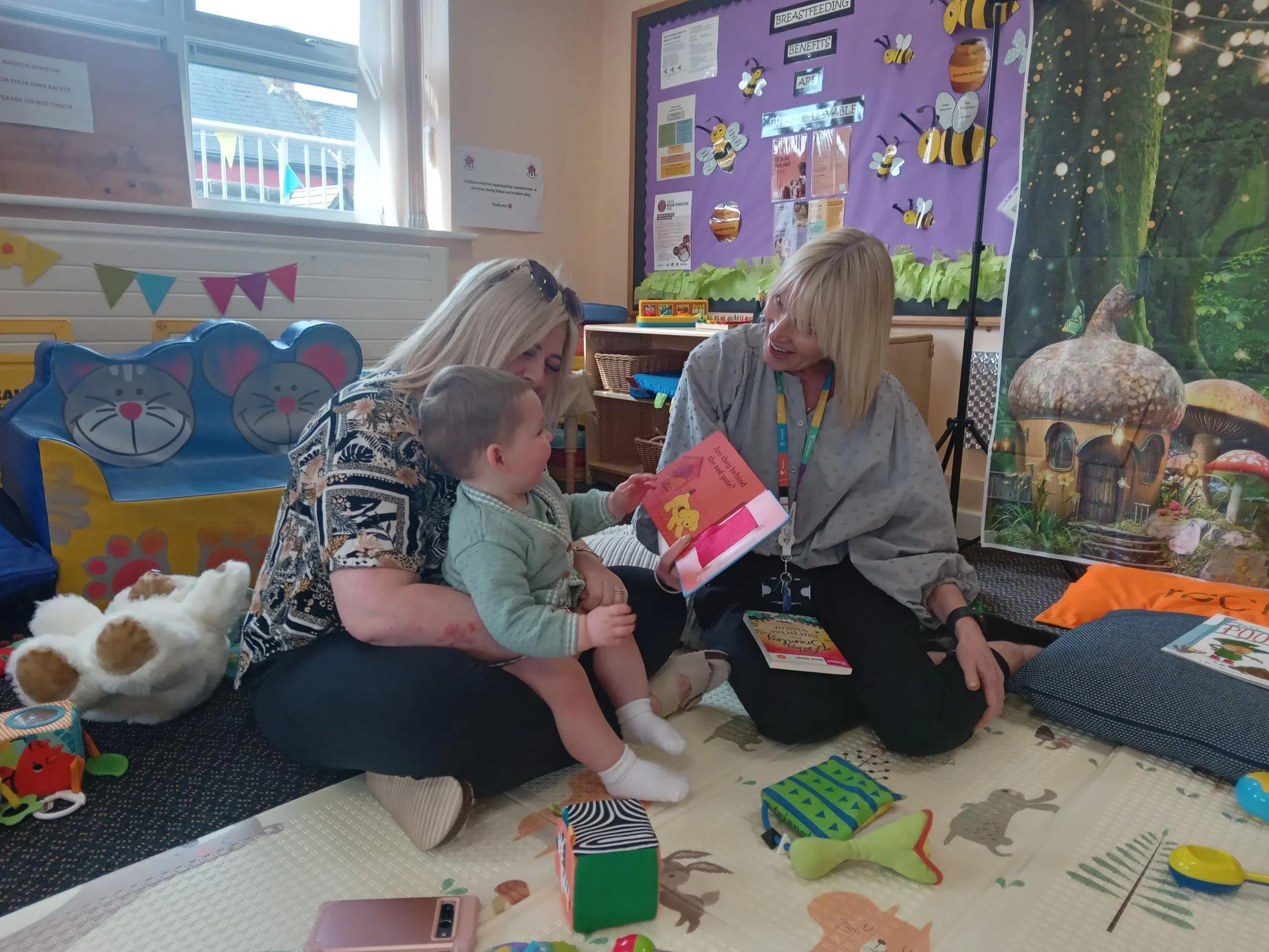 A woman holding a young child while another woman is reading a children's book to them in a colorful classroom with toys, stuffed animals, and a large fairy tale illustration in the background.