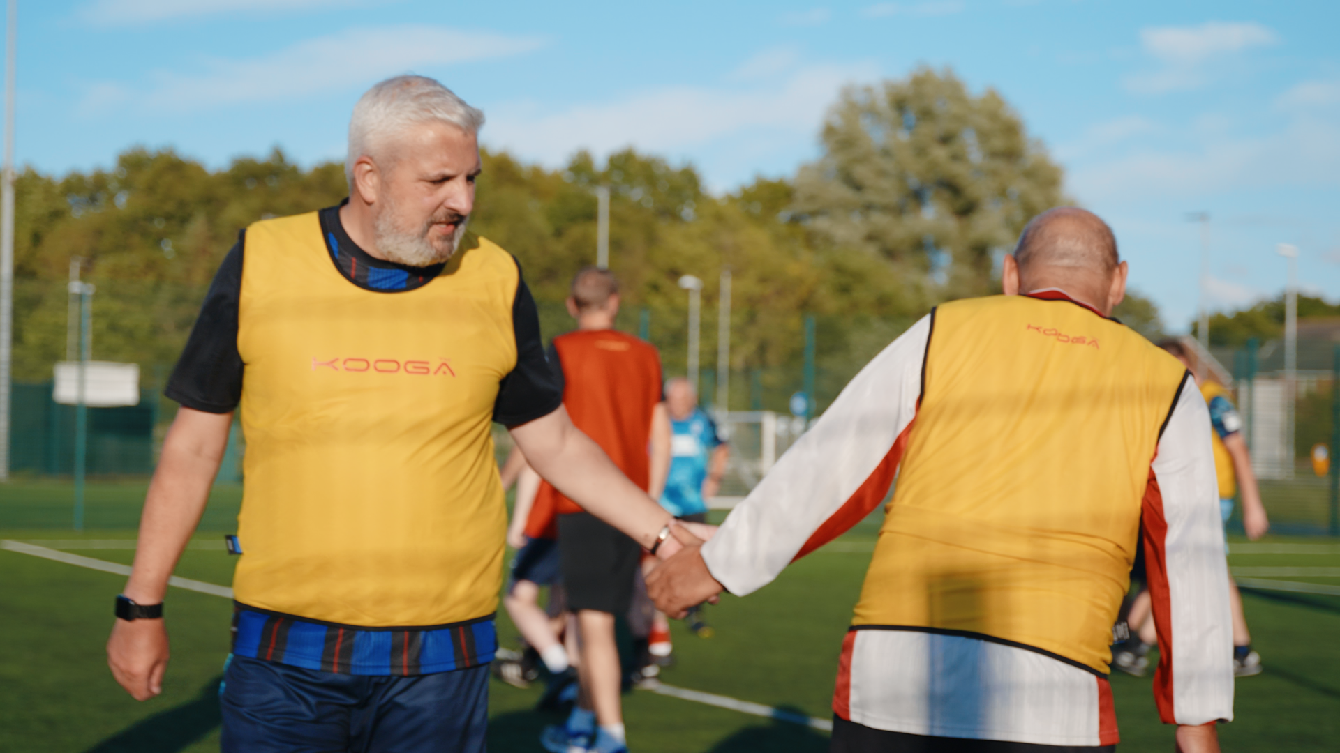 Two elderly men wearing yellow bibs and sports clothing, holding hands on a soccer field during a game or practice.