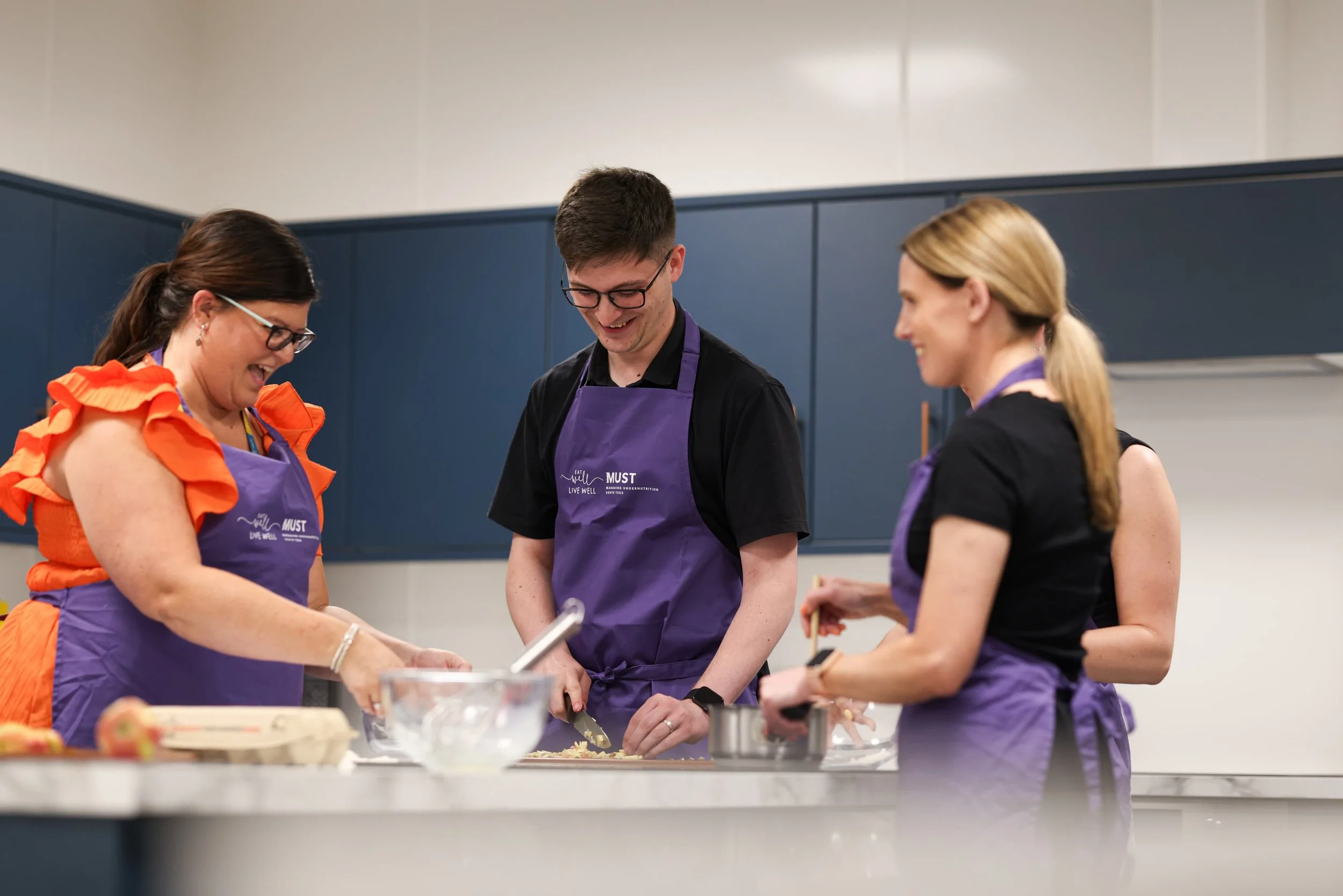 Three people cooking in a kitchen, two women and one man, all wearing purple aprons and smiling.