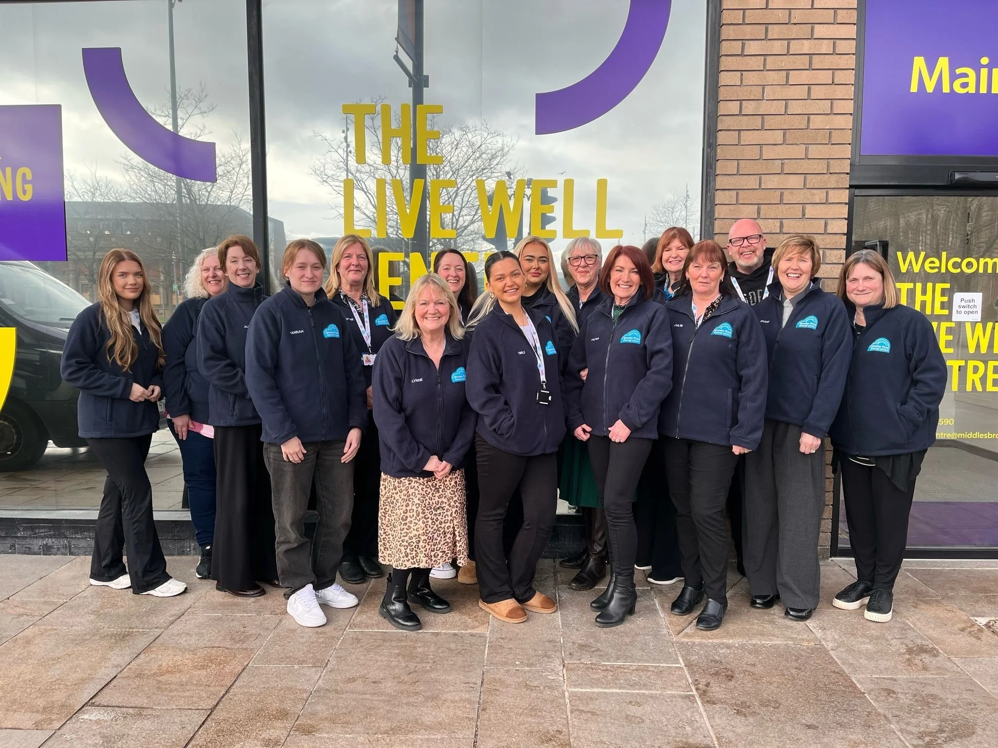 Group of diverse people in navy jackets standing outside a building with a glass front, smiling for the photo.