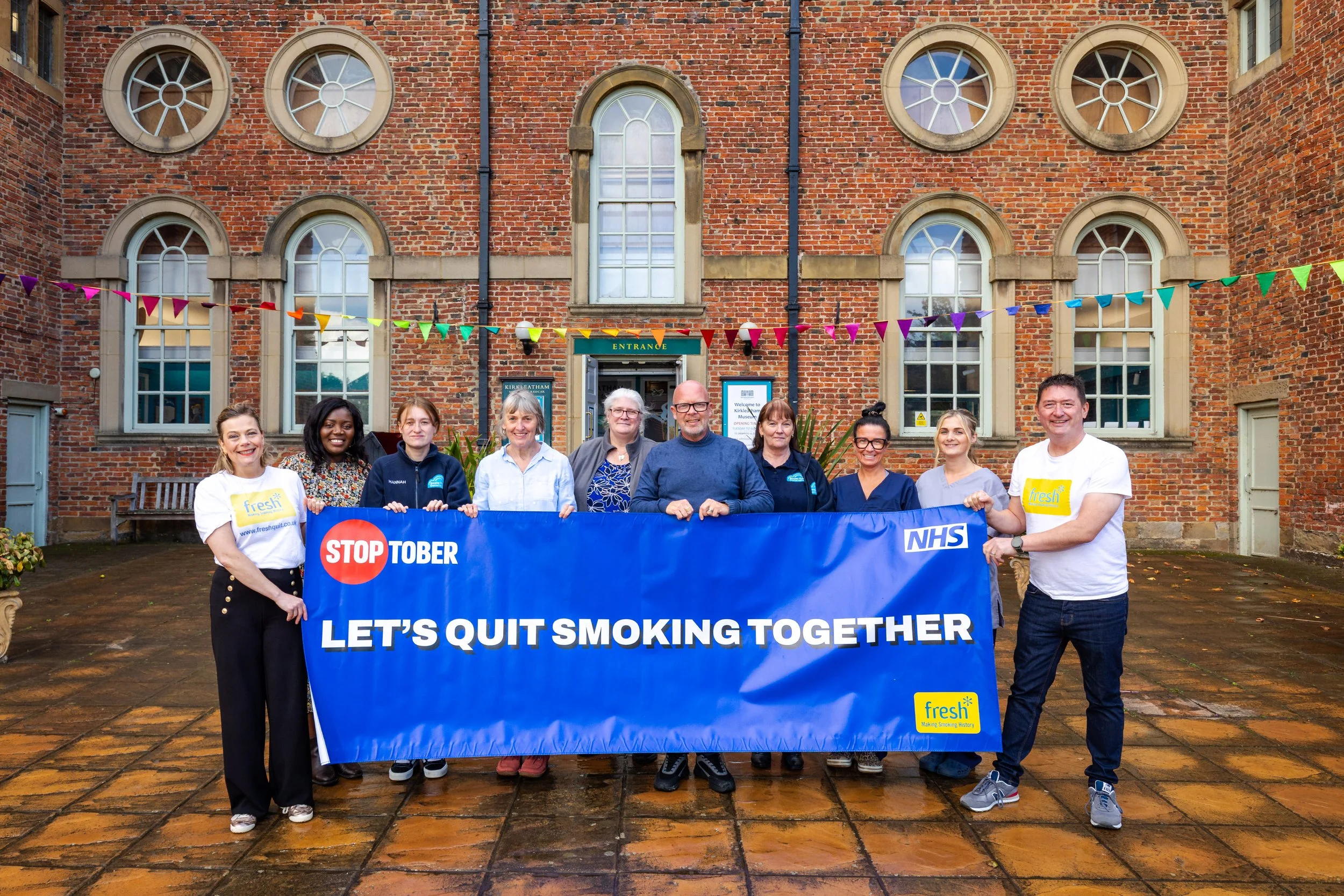 Group of diverse people holding a blue banner that says 'STOP TOBER, LET'S QUIT SMOKING TOGETHER' in front of a brick building with large windows and colorful bunting.