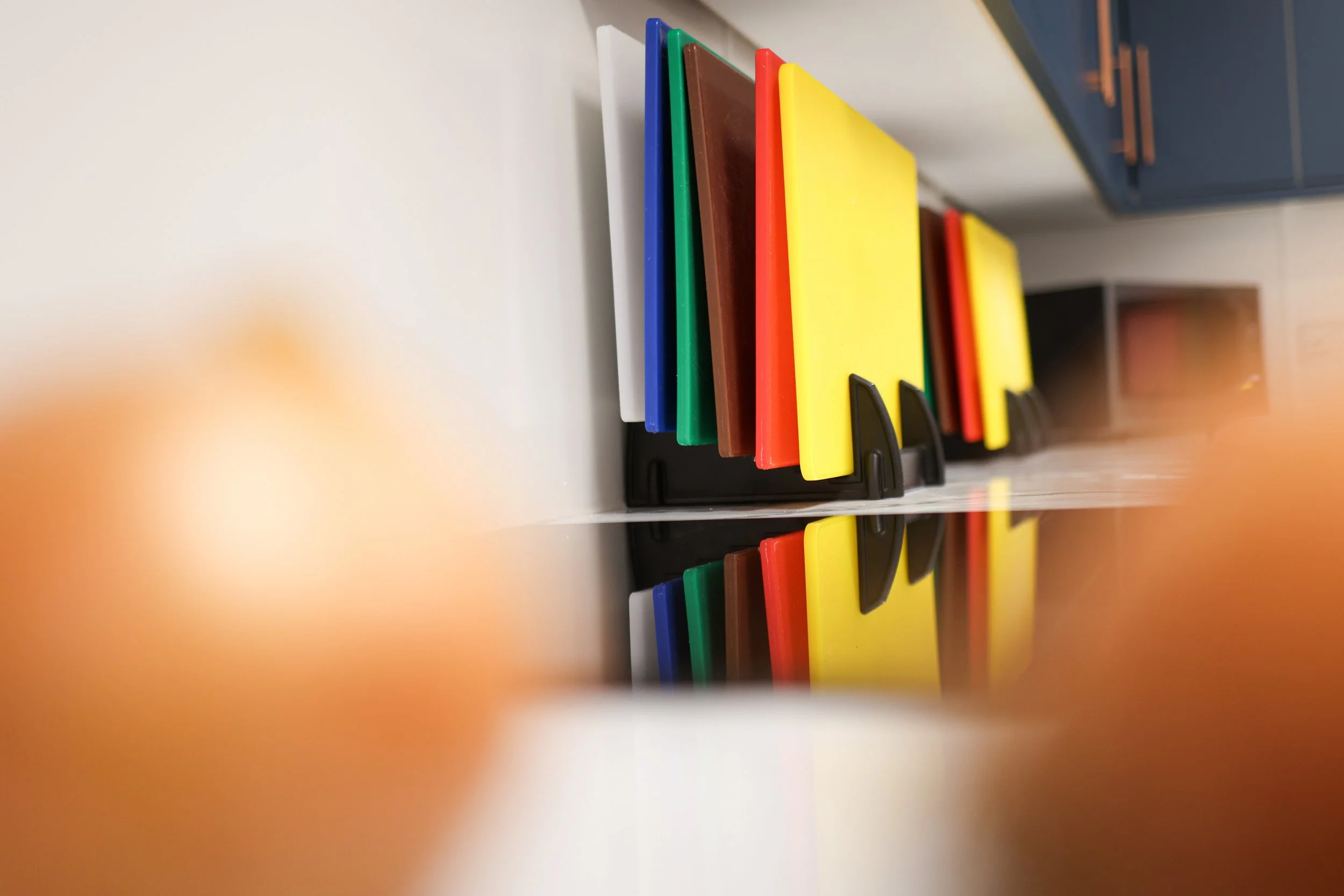 Colorful plastic cutting boards in white, blue, green, brown, red, and yellow, standing on black holders on a white countertop with a mirror reflecting the boards.