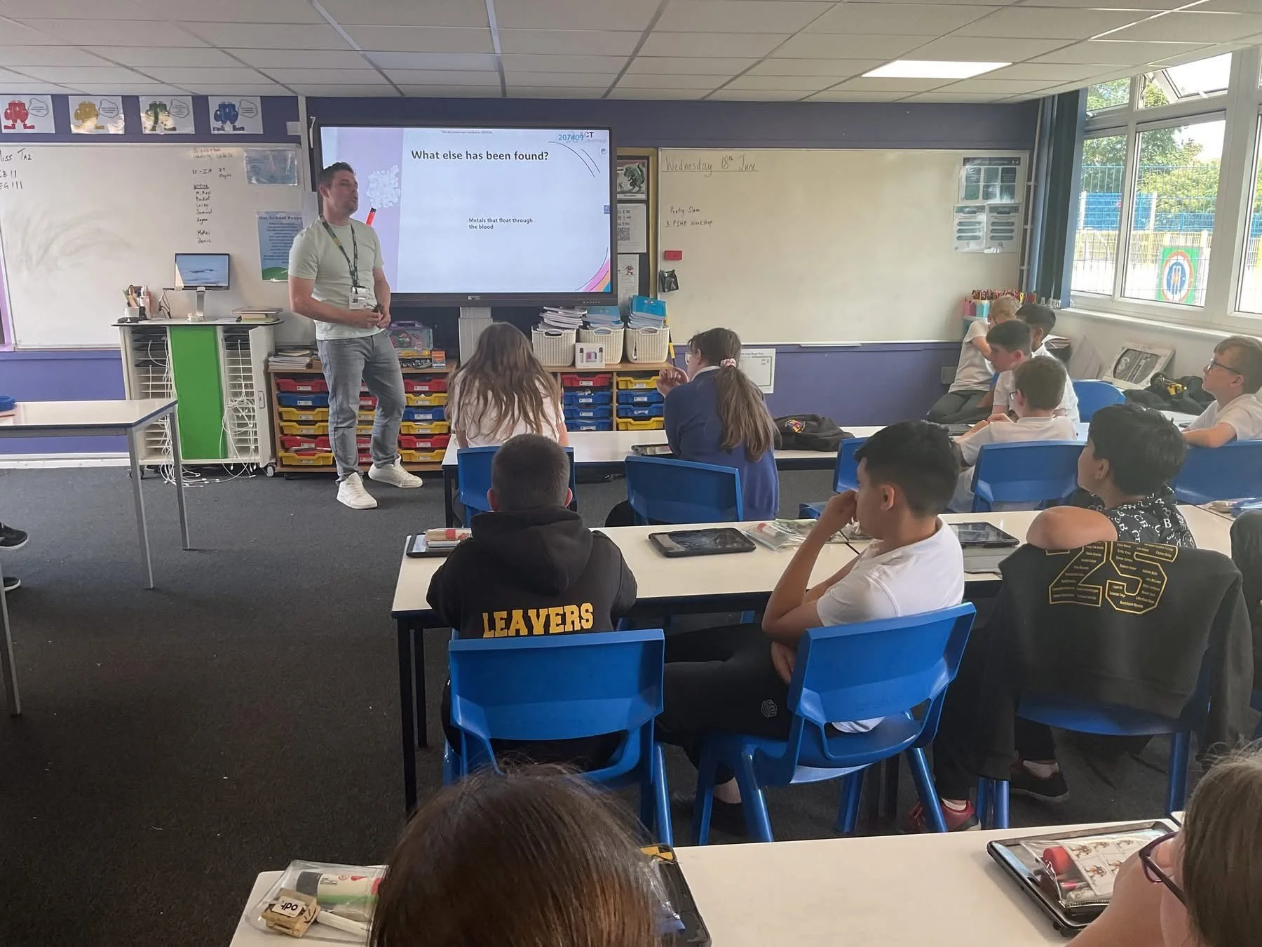A classroom with students seated at desks and a teacher standing in front of a digital whiteboard.