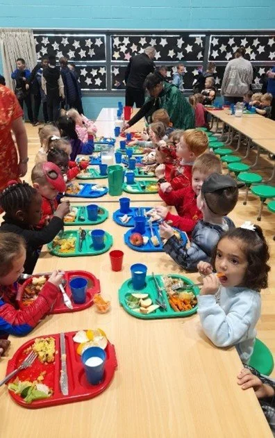 Children sitting at a long table with colorful trays and cups, enjoying a meal or snack in a decorated room with black and white star decorations on the window.