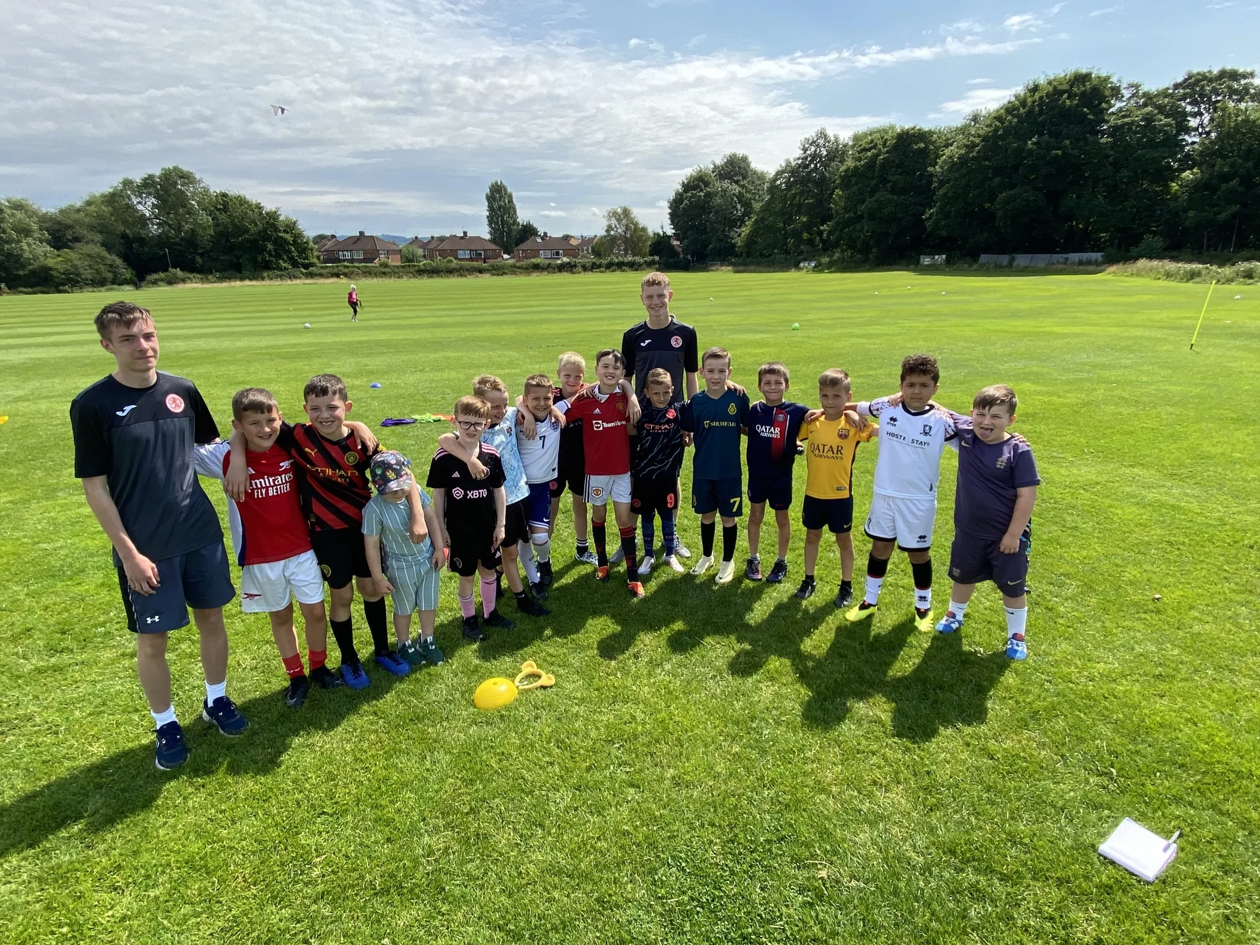 Group of children and two adult coaches standing together on a grassy field on a sunny day, wearing sports jerseys and shorts, with some sports equipment on the ground, and a partly cloudy sky overhead.