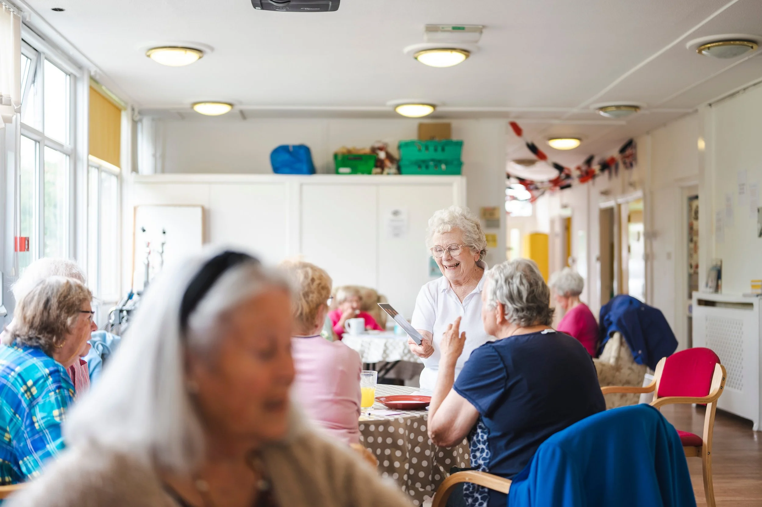 Elderly women gathered in a sunny room, chatting and smiling, with one woman standing and holding a tablet.