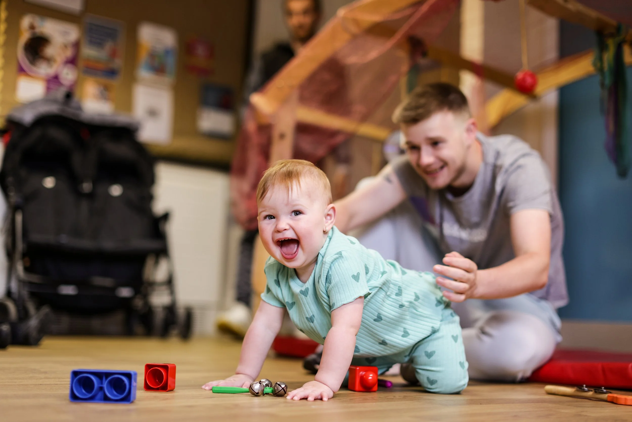 A young child crawling on a wooden floor, smiling and playing with toys, with an adult smiling behind them in a room decorated for a celebration.