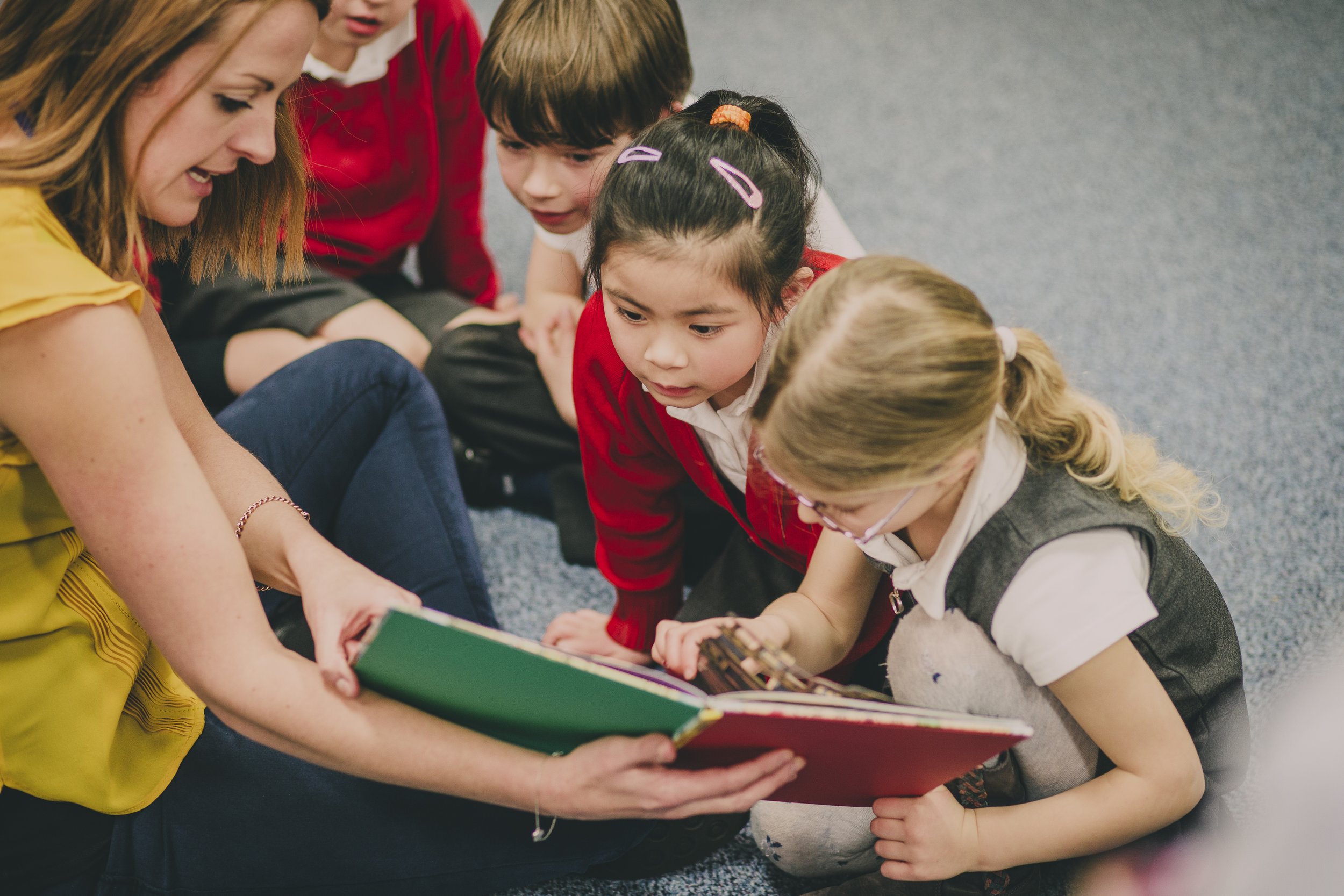 A female teacher reading a storybook to a group of children sitting on a carpeted floor.