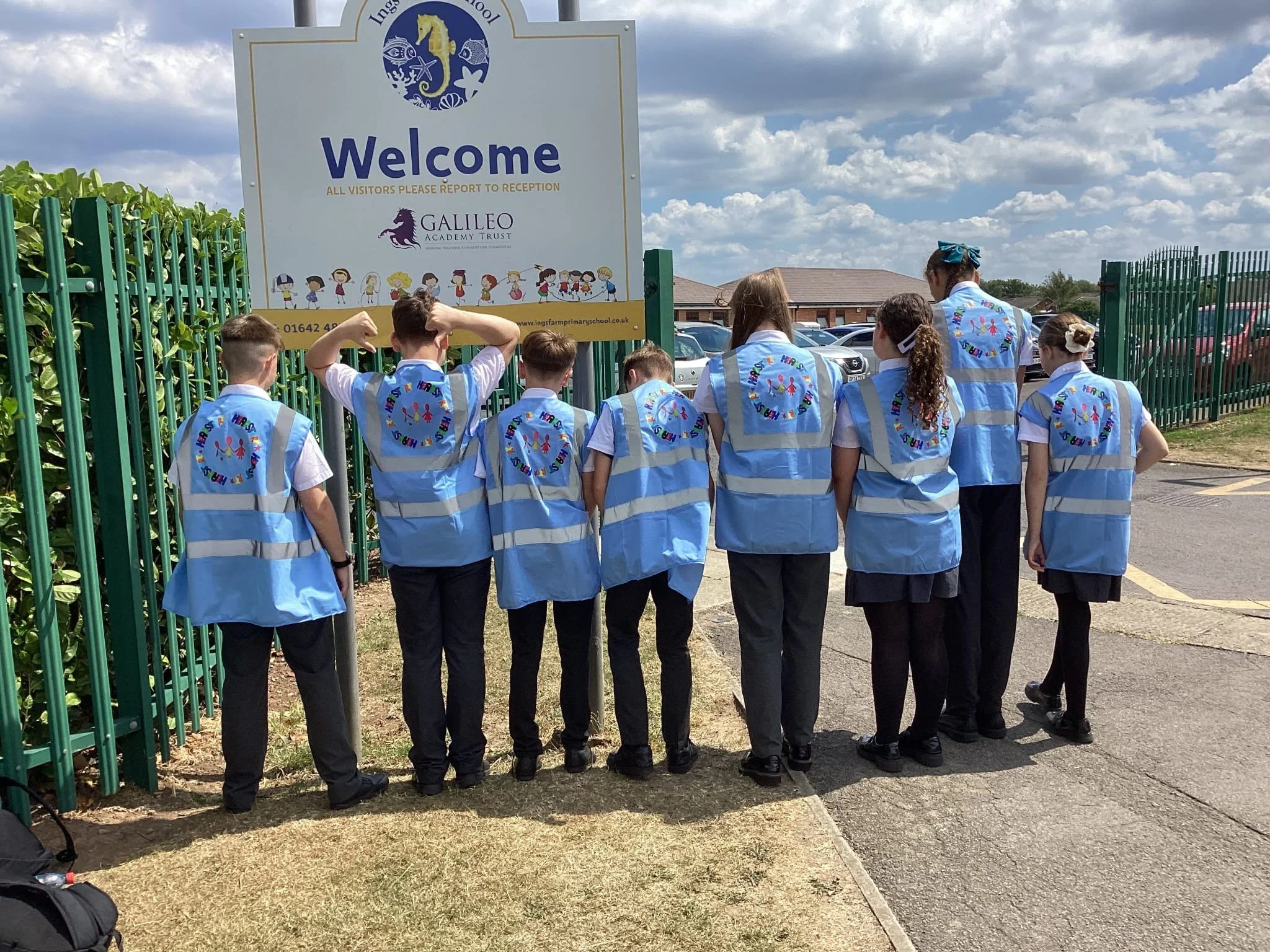 School children wearing in-school uniform and blue reflective safety vests, facing away from camera, standing near a school entrance gate with a welcome sign that reads "Welcome" and has a logo and cartoon children, on a partly cloudy day.