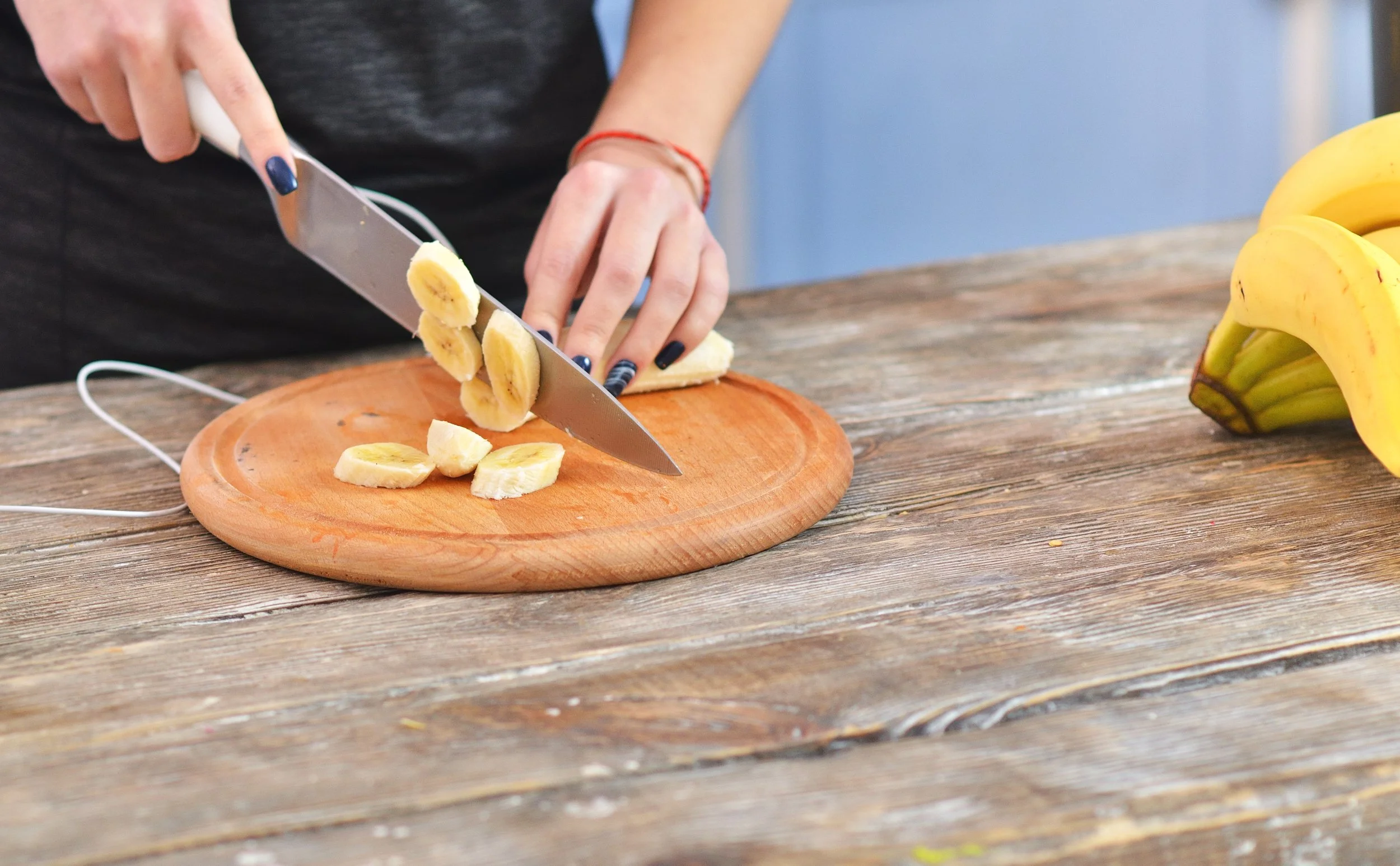 Person slicing a banana on a round wooden cutting board with a knife, with a bunch of bananas on a wooden table nearby.