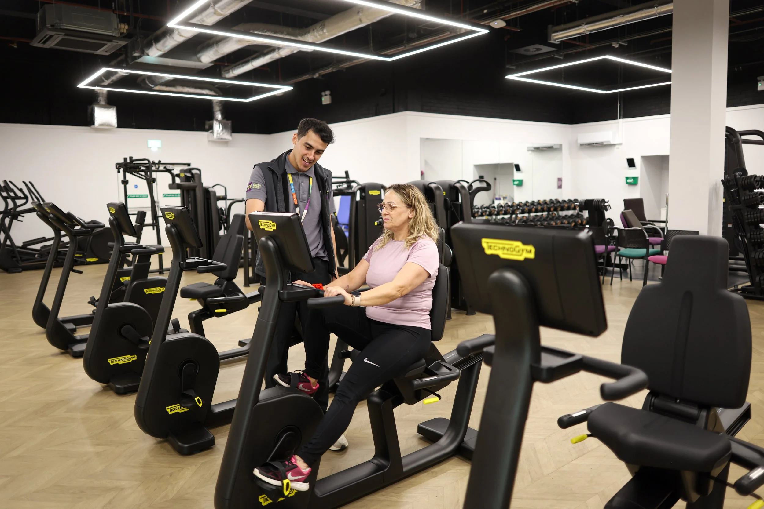 A woman at a gym on a stationary exercise bike, talking with a trainer, in a spacious fitness center filled with exercise machines and equipment.