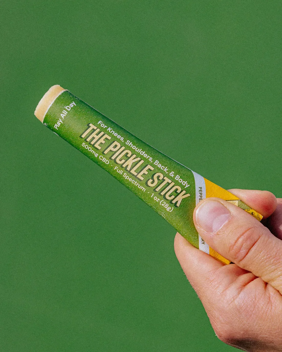 A person holding a tube of 'The Pickle Stick,' a CBD balm for knees, shoulders, back, and body against a green background.