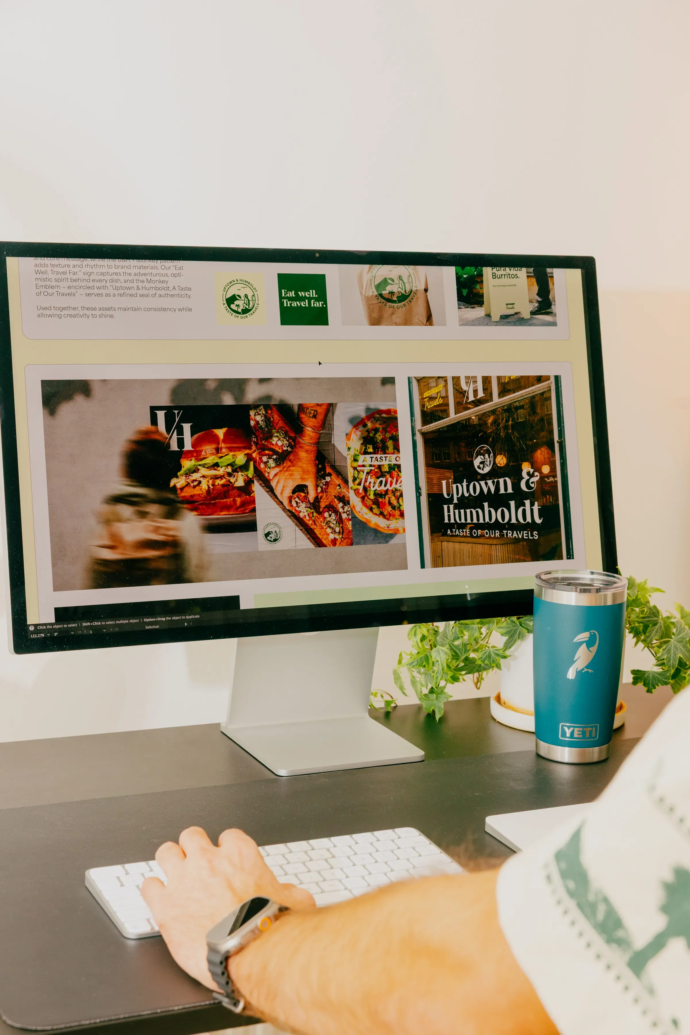 Computer monitor displaying a website for Uptown & Humboldt with images of food, a person blurred moving, and signs. Desk with a white keyboard, a person's arm, a YETI tumbler, and some green plants.