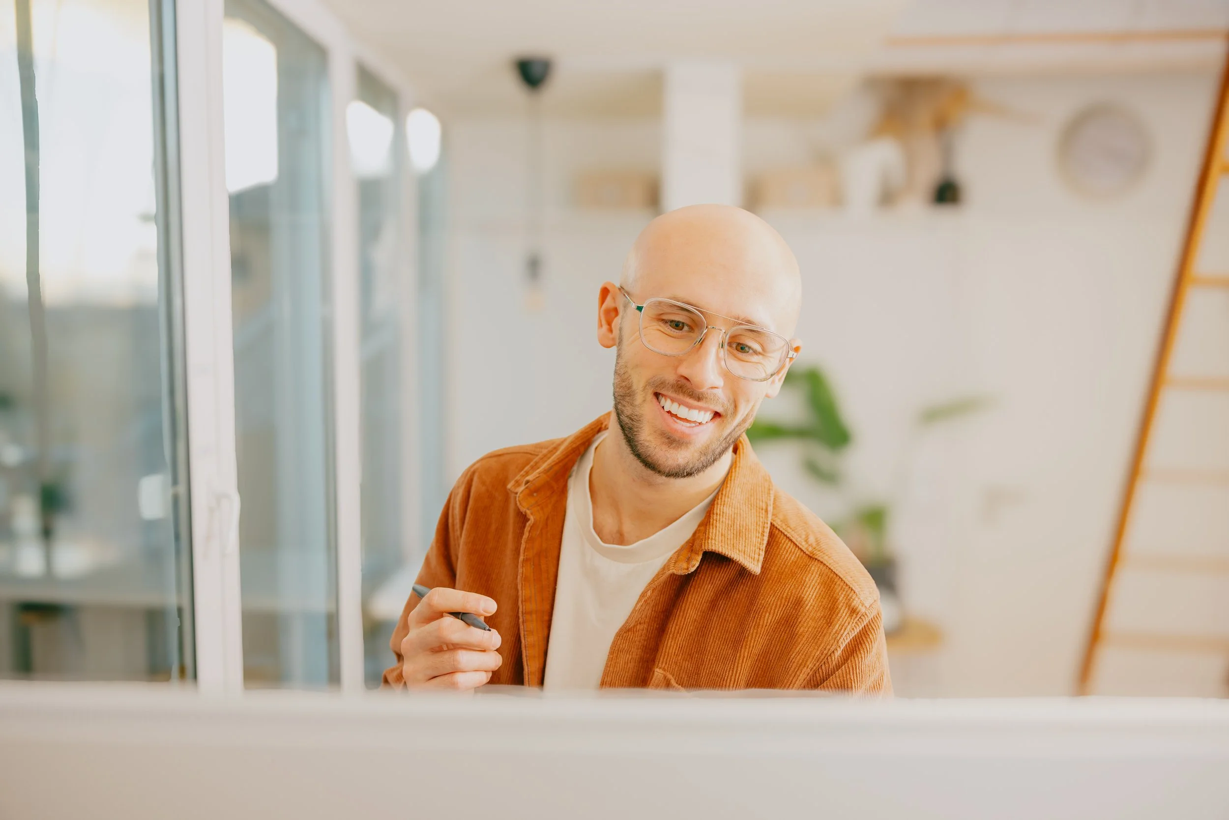 A smiling bald man with glasses wearing a burnt orange shirt and white t-shirt, holding a stylus, sitting at a desk in a bright modern home or office space.