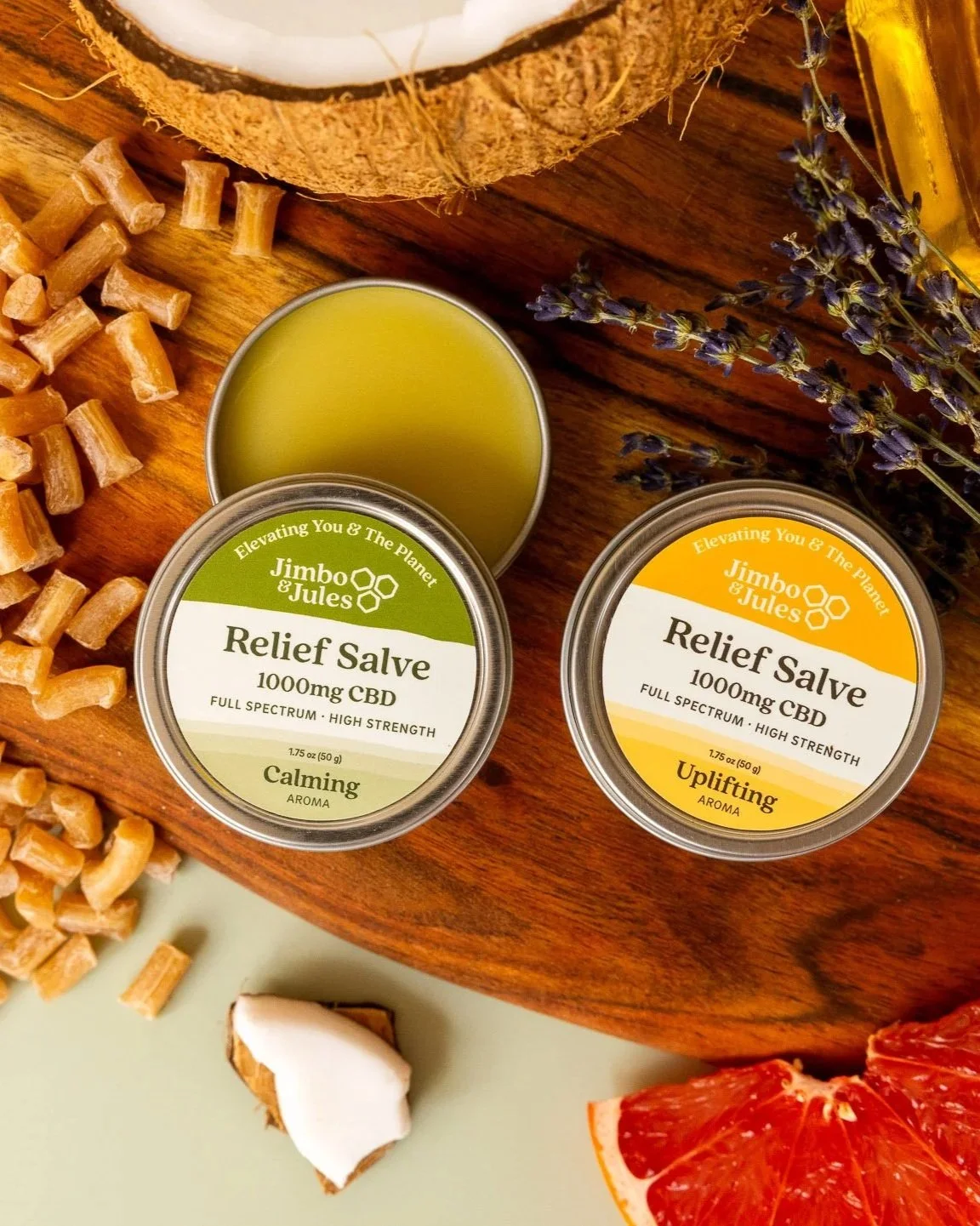 Two jars of Jimbo & Jules relief salve with CBD, one labeled calming and the other uplifting, placed on a wooden surface alongside a small bowl of green balm, dried herbs, a piece of coconut, pasta, and slices of grapefruit.