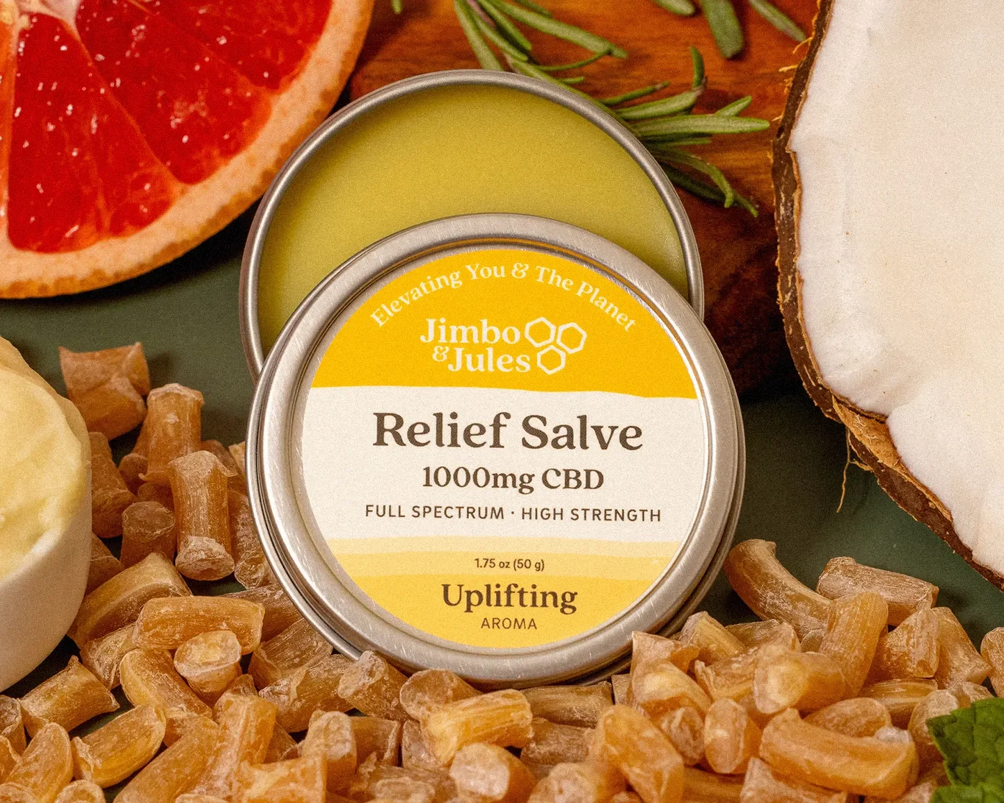 A tin container of Jimbo & Jules Relief Salve with 1000mg CBD, surrounded by various natural ingredients including a grapefruit slice, green leaves, coconut, and chunks of raw amber-colored crystals.