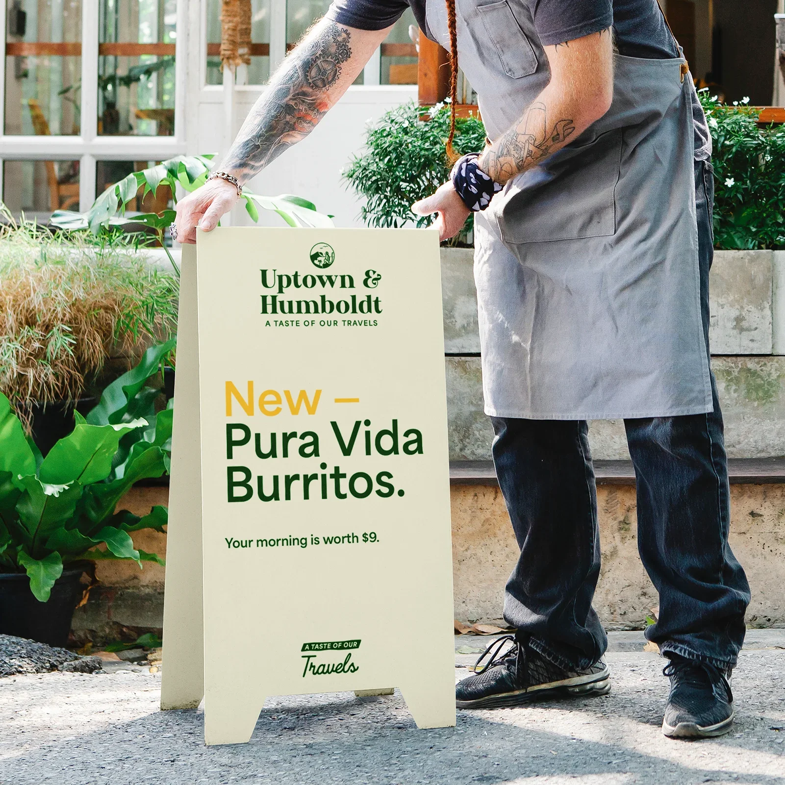 A person adjusting a signboard outdoors with plants and a building in the background. The signboard advertises New Pura Vida Burritos from Uptown & Humboldt, highlighting the value of morning meals at $9.